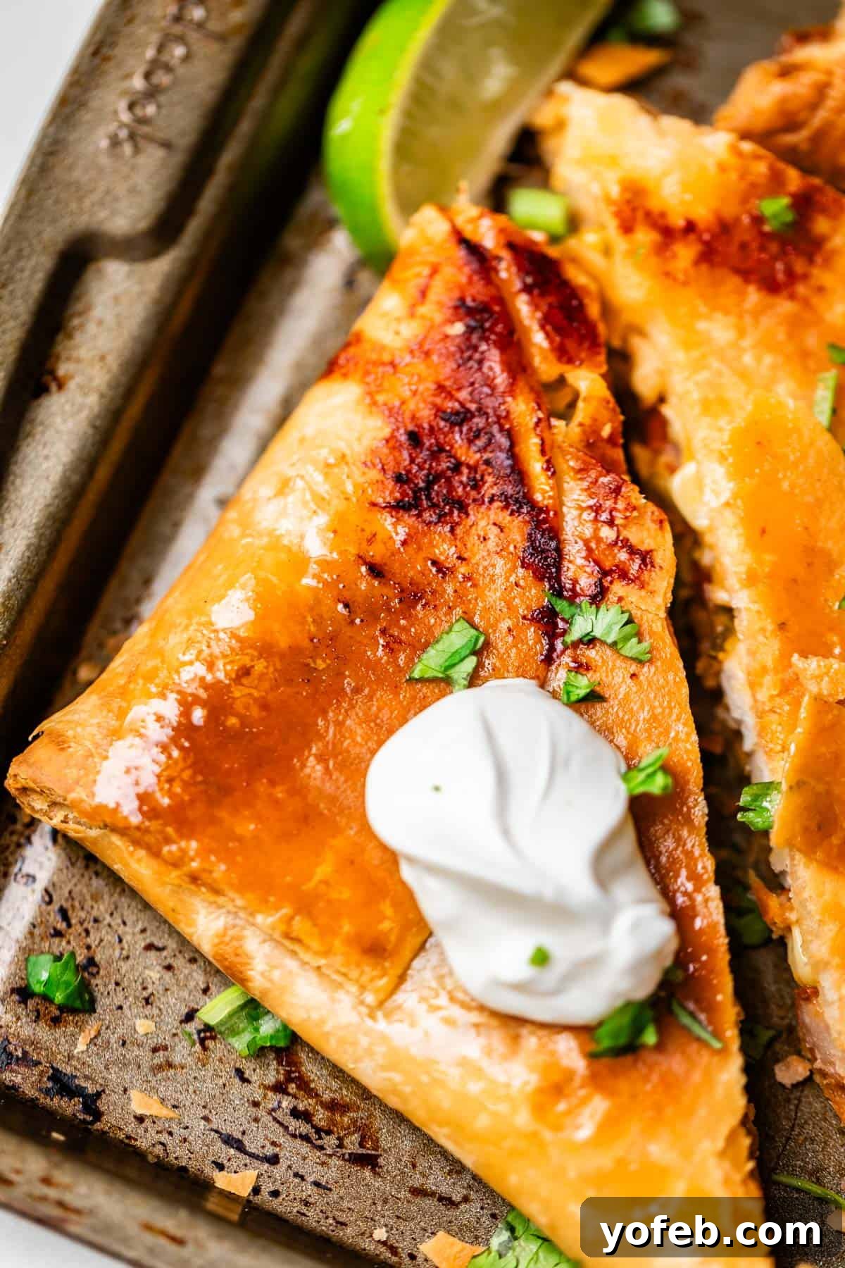 Crispy Sheet Pan Chicken Quesadillas 6 A close-up of a golden, crispy quesadilla slice topped with a dollop of sour cream and chopped cilantro, served on a baking tray with a lime wedge in the background.