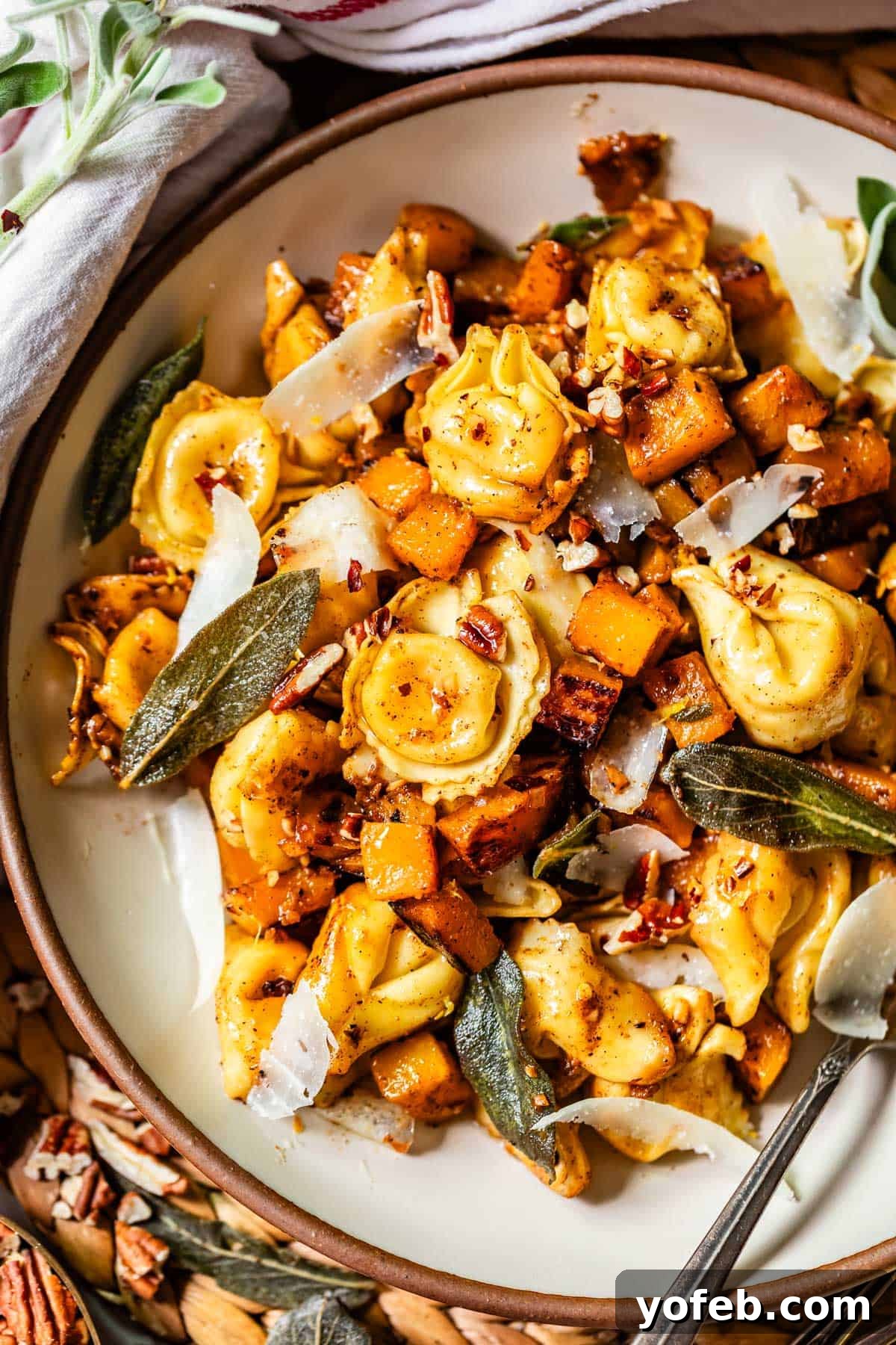 Butternut Bliss Tortellini 2 A plate of cheese tortellini mixed with roasted butternut squash, crispy sage leaves, shaved parmesan, pecans, and spices, served with a fork. The dish has a golden, autumn-inspired appearance.
