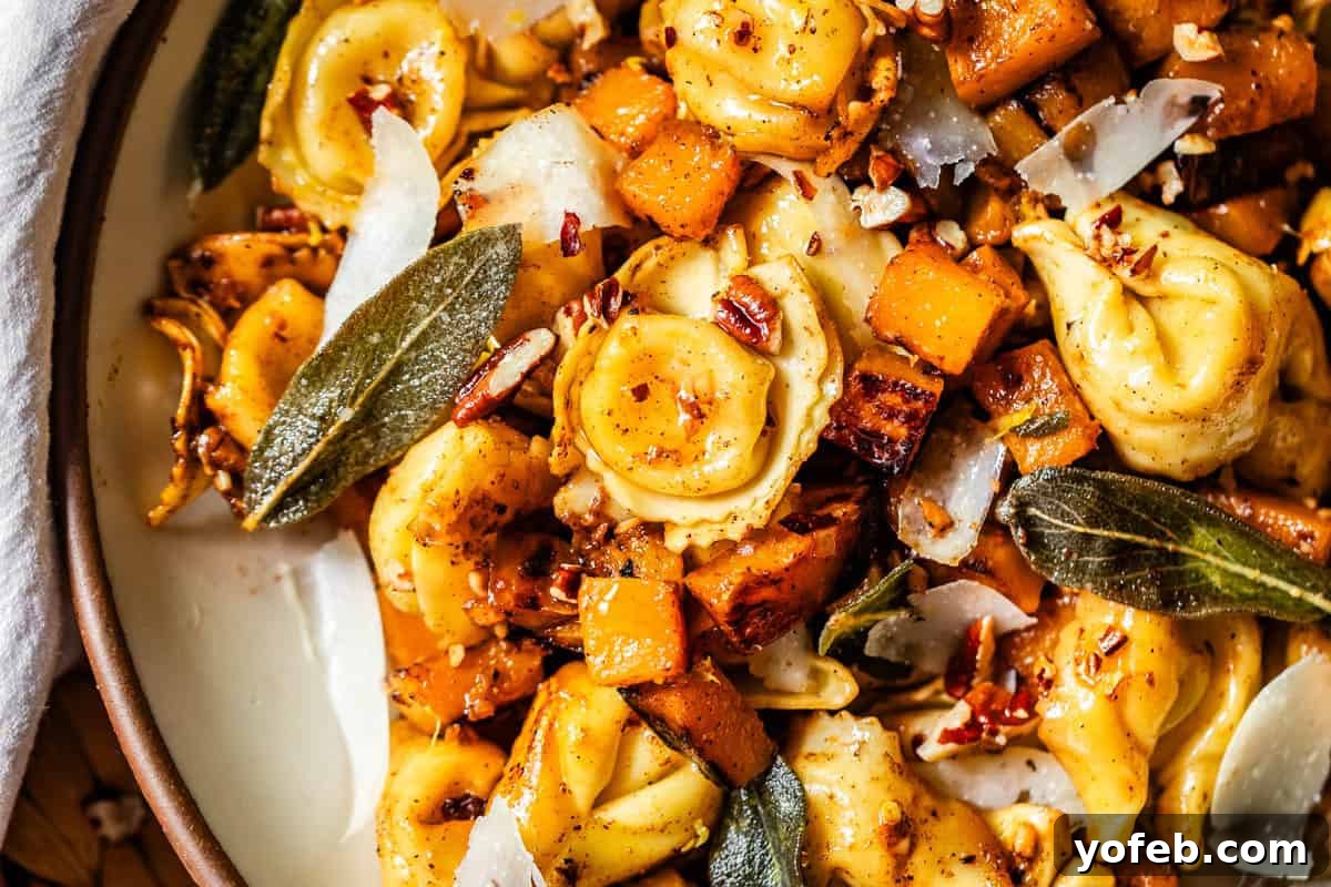Butternut Bliss Tortellini 17 A close-up of a dish featuring cheese tortellini, roasted butternut squash, crispy sage leaves, pecans, and shaved Parmesan on a white plate. The ingredients are golden and garnished with herbs.