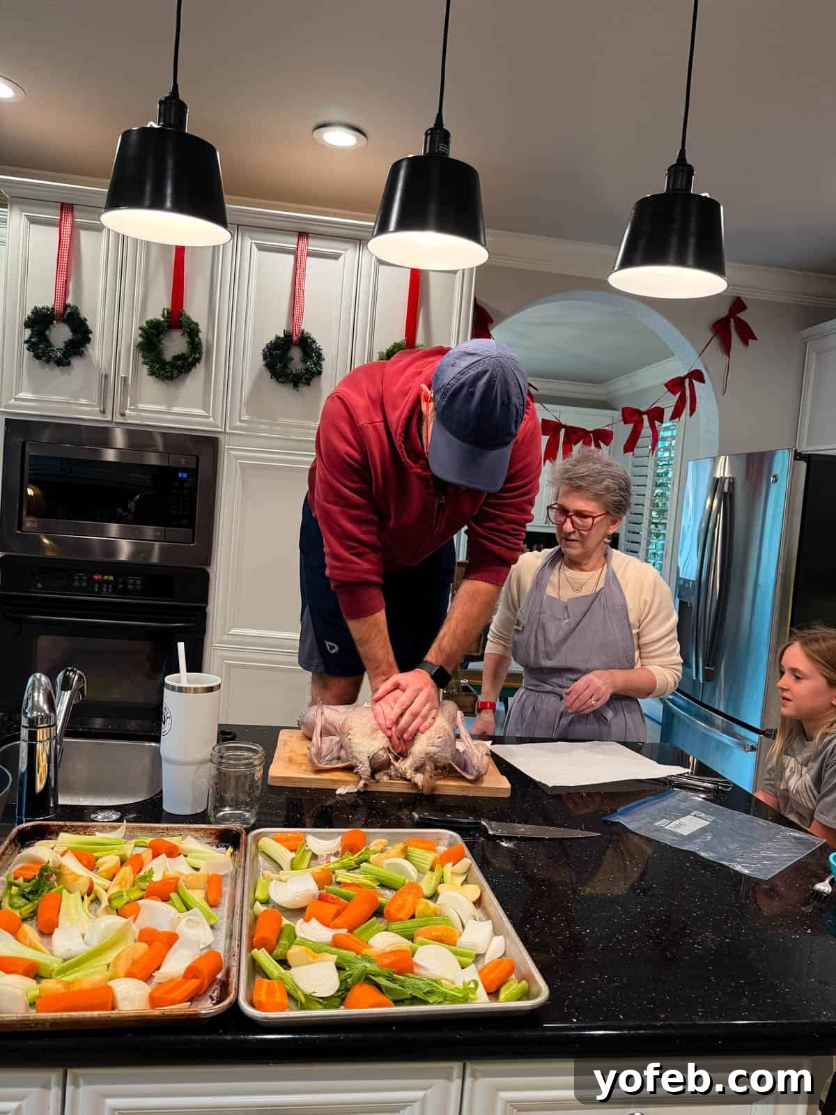 Butternut Bliss Tortellini 3 A man in a red hoodie stands on a kitchen counter, preparing a turkey while a woman and a girl watch. Trays of chopped vegetables are on the counter, and the kitchen is decorated for the holidays.