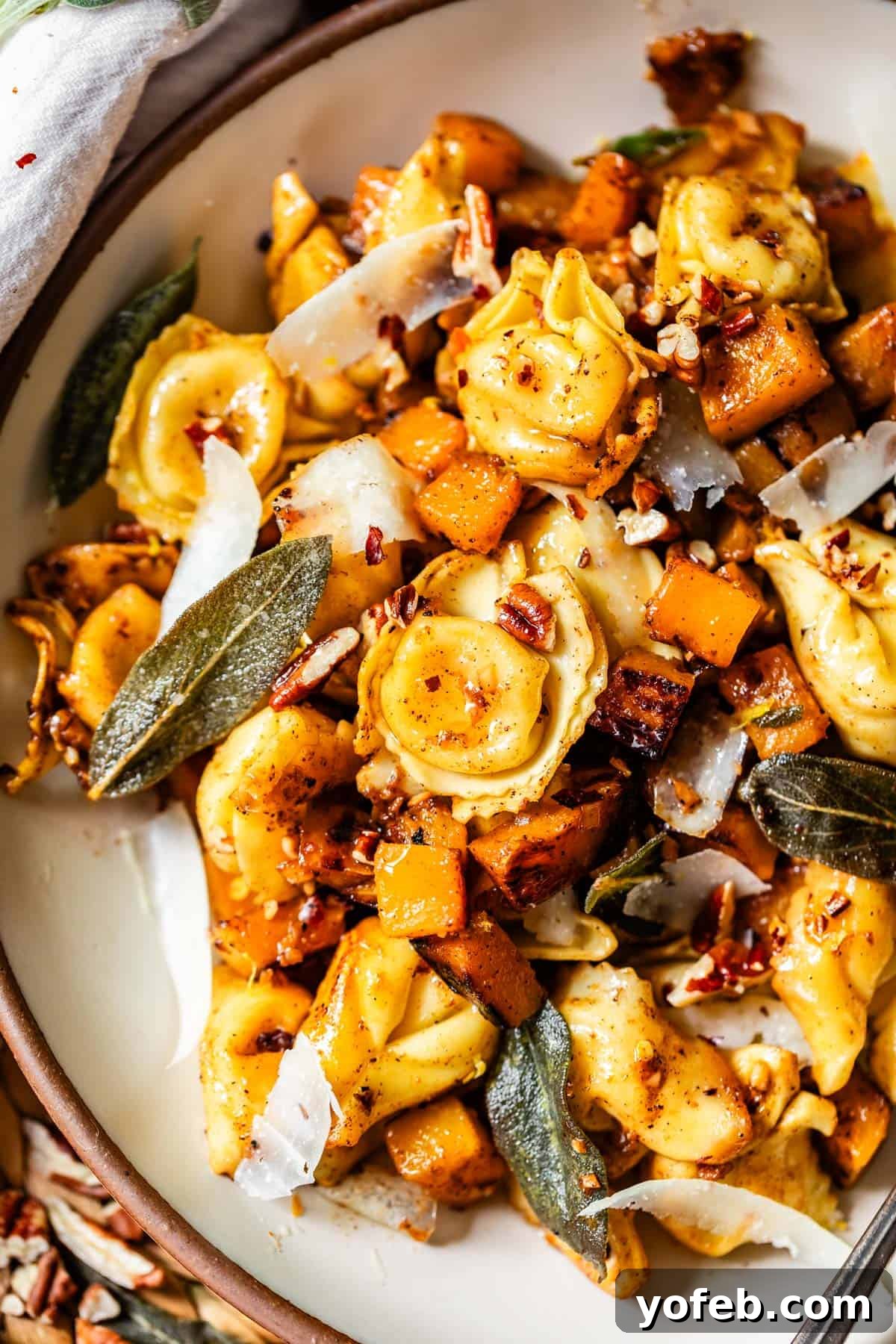 Butternut Bliss Tortellini 20 A close-up of a pasta dish featuring cheese-filled tortellini, roasted butternut squash, fresh sage leaves, shaved parmesan, and chopped nuts on a white plate. The meal looks vibrant and garnished with herbs.