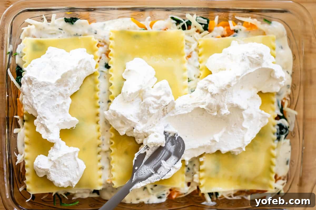 Creamy Golden Butternut Lasagna 22 A close-up of a baking dish filled with uncooked lasagna. A spatula spreads a thick layer of ricotta cheese over pasta sheets, with visible spinach, sauce, and shredded cheese underneath.