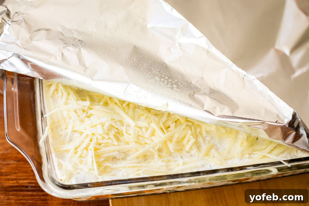 Creamy Golden Butternut Lasagna 24 A glass baking dish filled with shredded cheese and a creamy mixture, partially covered with a sheet of aluminum foil, sits on a wooden surface.