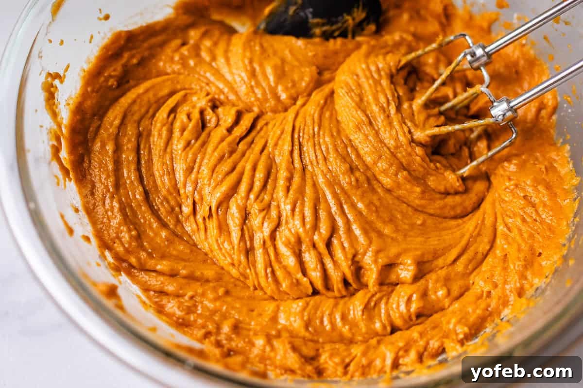 Autumn Spiced Pumpkin Sheet Cake with Maple Cream Cheese Frosting 12 Close-up of a glass mixing bowl filled with thick, orange pumpkin batter being mixed with an electric hand mixer, showing the smooth, final consistency.