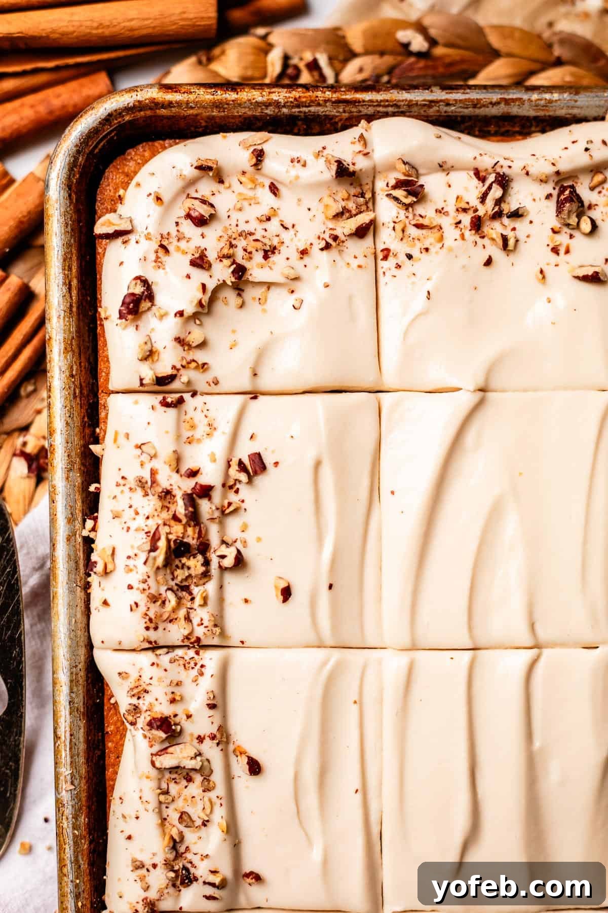 Autumn Spiced Pumpkin Sheet Cake with Maple Cream Cheese Frosting 18 A close-up of a frosted sheet cake cut into squares, topped with chopped pecans. Swirls in the creamy frosting are visible, and cinnamon sticks surround the cake on the table, presenting a delightful and ready-to-serve dessert.