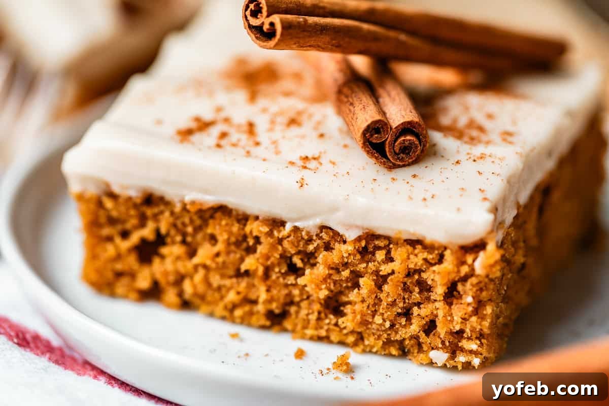 Autumn Spiced Pumpkin Sheet Cake with Maple Cream Cheese Frosting 19 A close-up of a square piece of spiced cake with white frosting, sprinkled with cinnamon and topped with two cinnamon sticks, served on a white plate, showcasing its festive presentation.