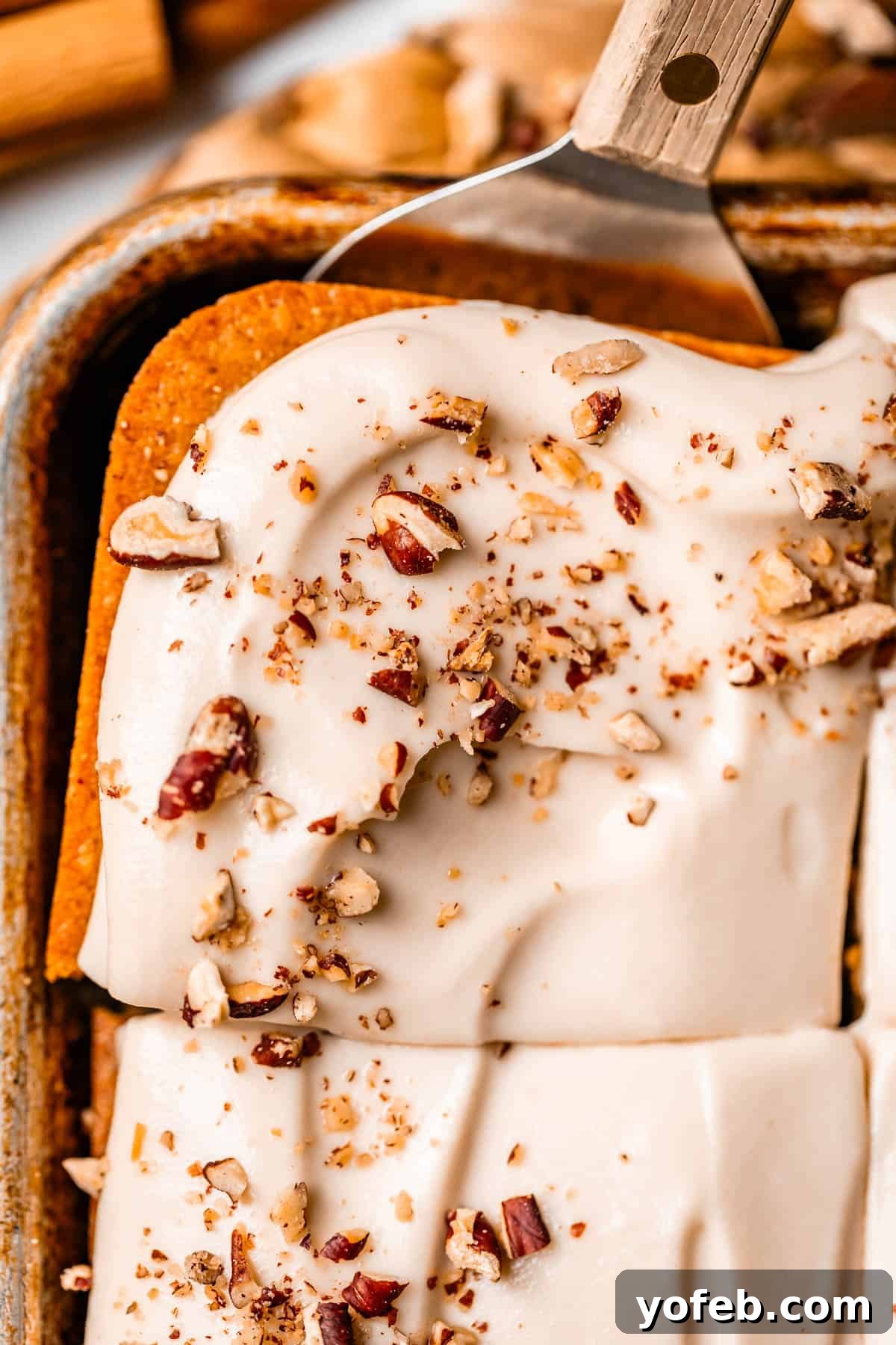 Autumn Spiced Pumpkin Sheet Cake with Maple Cream Cheese Frosting 20 A spatula lifts a square piece of pumpkin sheet cake topped with creamy white frosting and sprinkled with chopped pecans from a baking pan, highlighting its perfect portion and tempting appeal.
