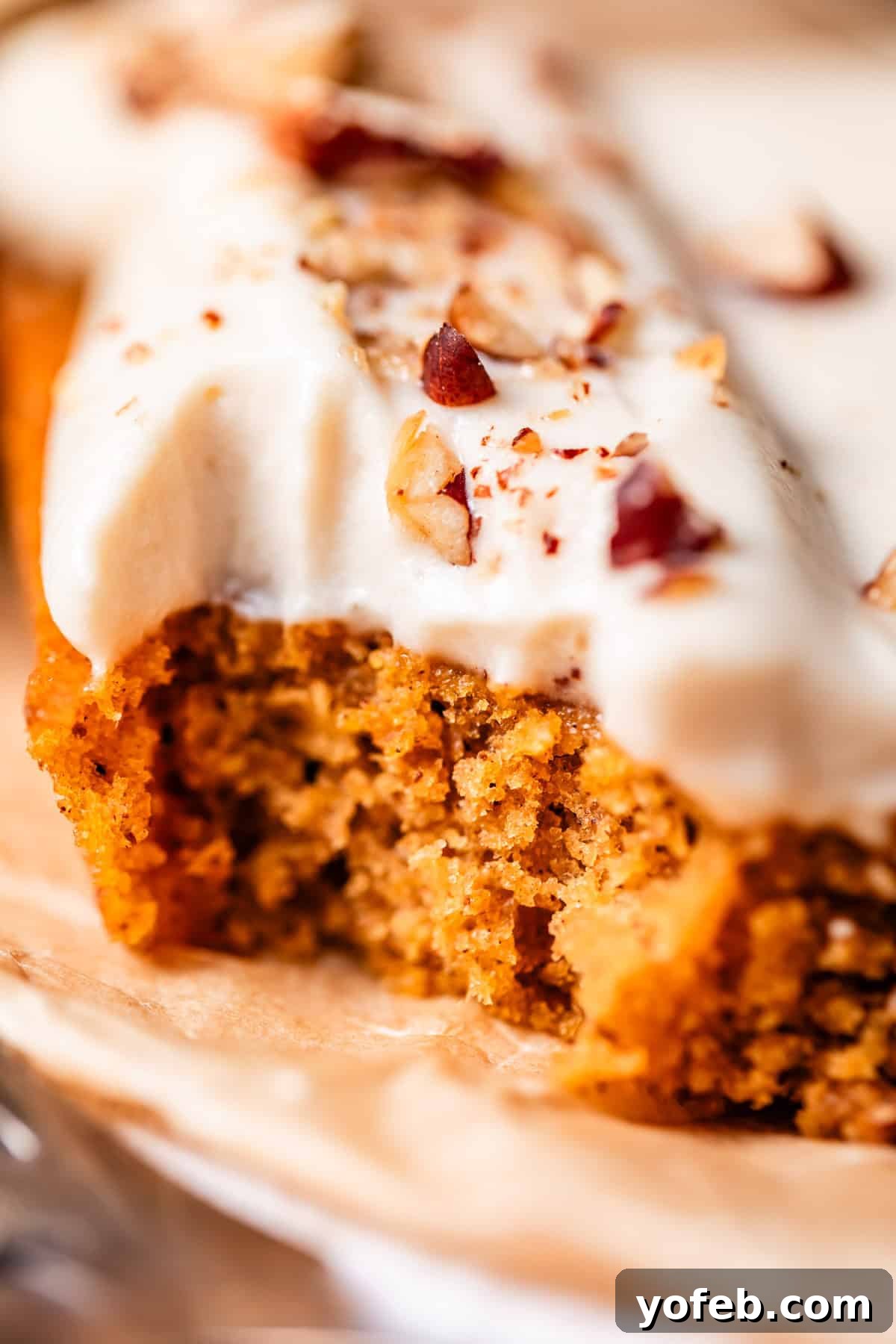 Autumn Spiced Pumpkin Sheet Cake with Maple Cream Cheese Frosting 4 A close-up of a slice of moist, spiced cake topped with creamy white frosting and sprinkled with chopped nuts, resting on a piece of parchment paper, highlighting its delicious texture.