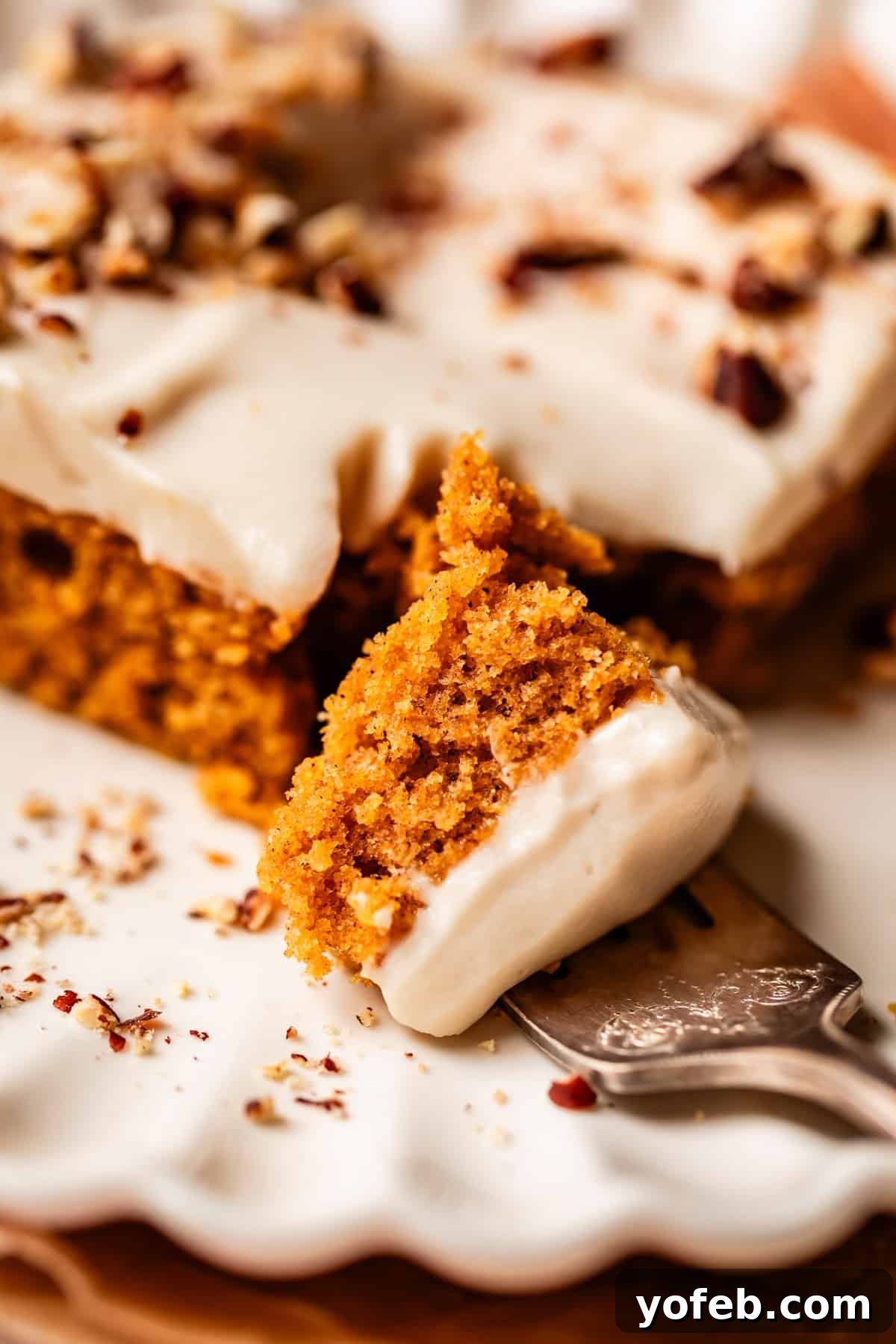 Autumn Spiced Pumpkin Sheet Cake with Maple Cream Cheese Frosting 5 A close-up of a moist slice of carrot cake topped with creamy frosting and chopped nuts, resting on a decorative plate with a fork holding a bite-sized piece, showcasing a rich and enticing dessert.