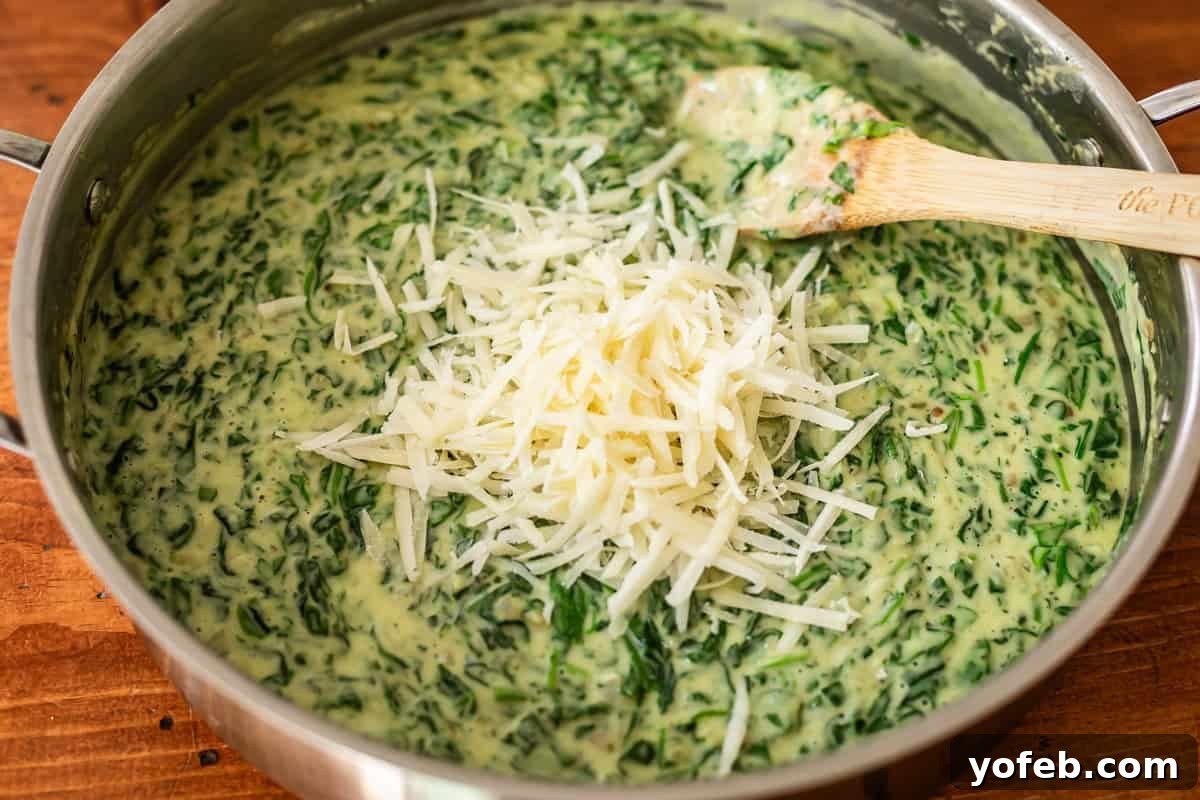 A pan filled with creamy spinach dip is topped with a pile of shredded cheese, and a wooden spoon rests inside the mixture. The pan sits on a wooden surface.