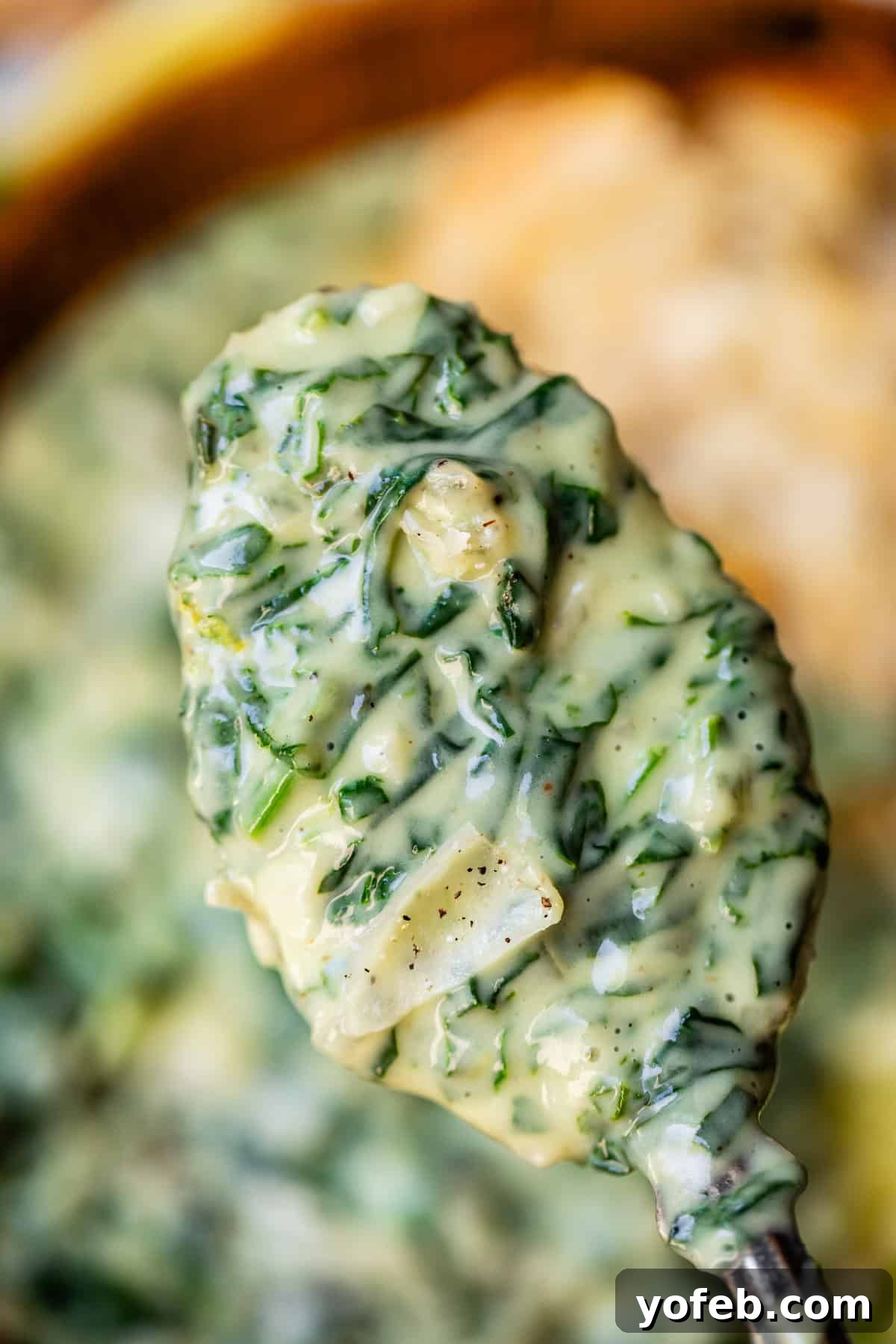 A close-up of a spoonful of creamy spinach artichoke dip, showing chopped spinach and bits of artichoke in a thick, cheesy sauce with visible seasoning flecks.
