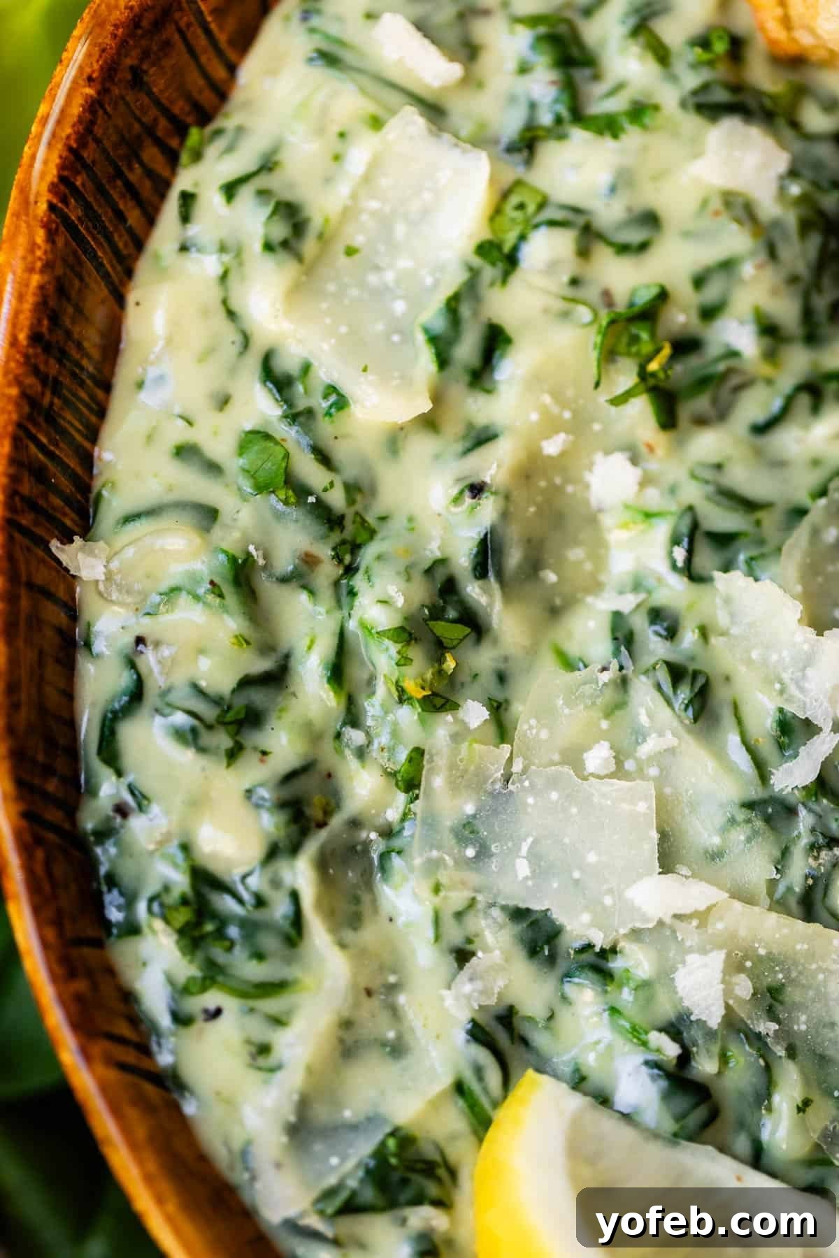 Close-up of creamy spinach dip in a wooden bowl, topped with shaved parmesan cheese and a lemon wedge. The dip has a rich, smooth texture with visible bits of spinach.