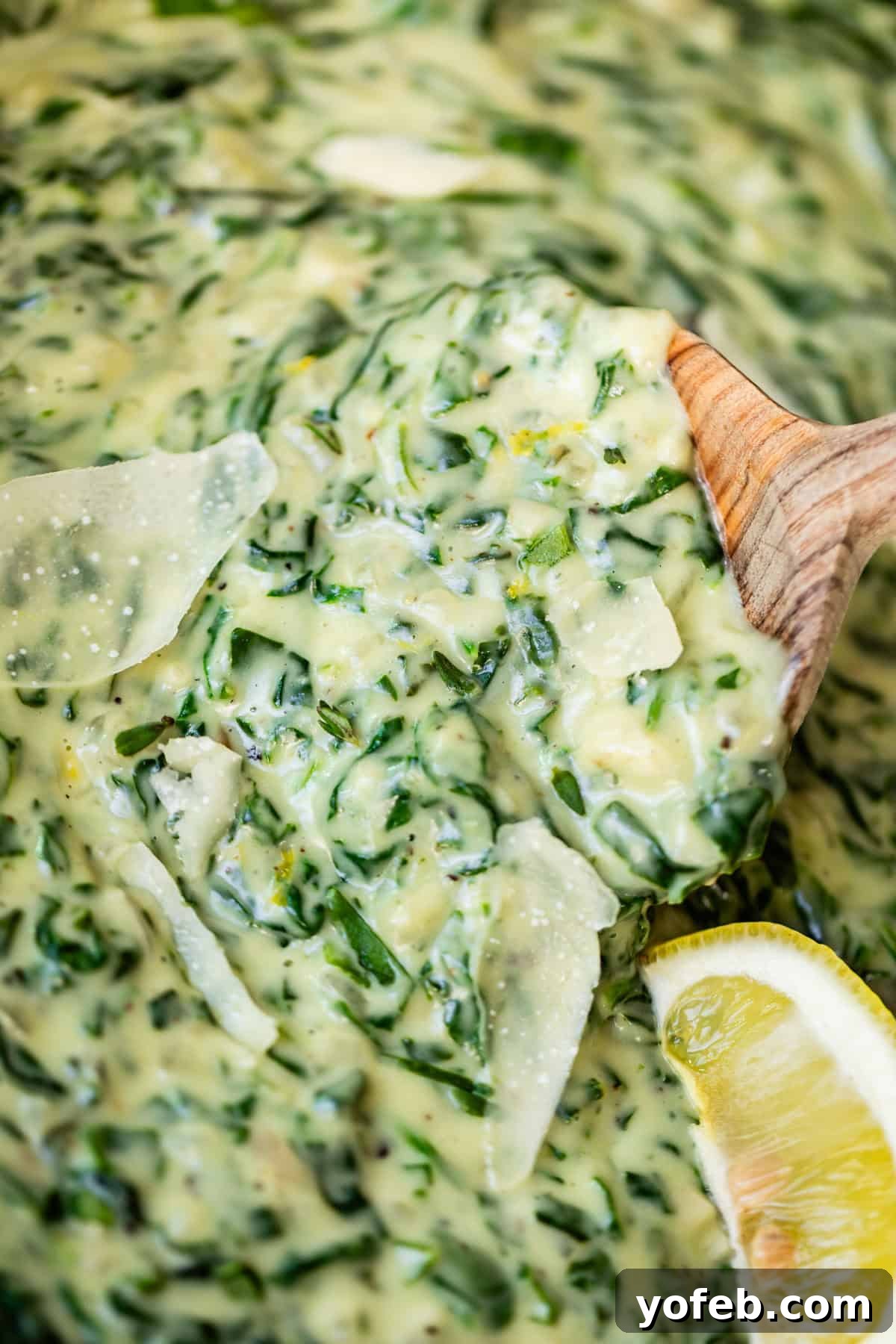 Close-up of creamy spinach dip with visible cheese shavings, held up by a wooden spoon. A wedge of lemon is positioned on the side of the dish.