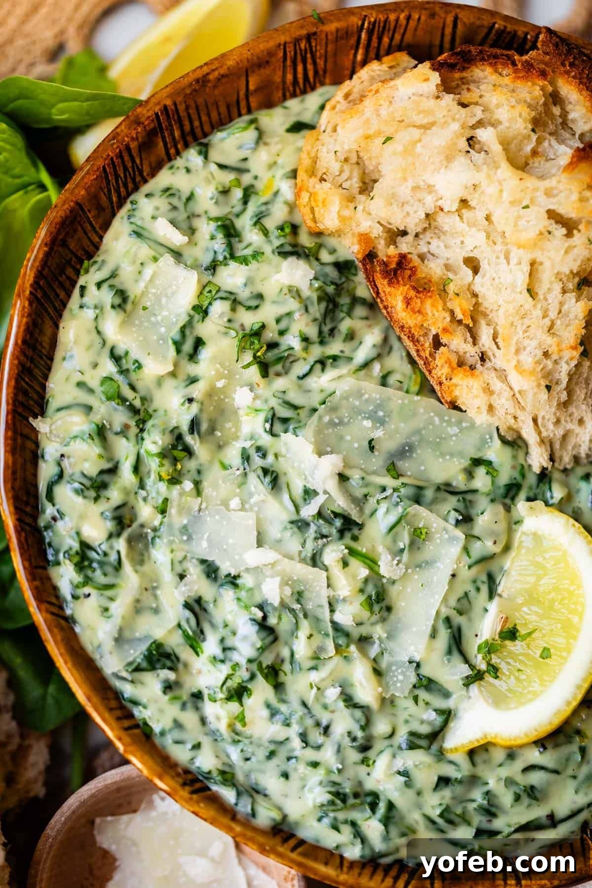 A wooden bowl filled with creamy spinach dip topped with Parmesan shavings, served with a slice of rustic bread and a wedge of lemon on the side.