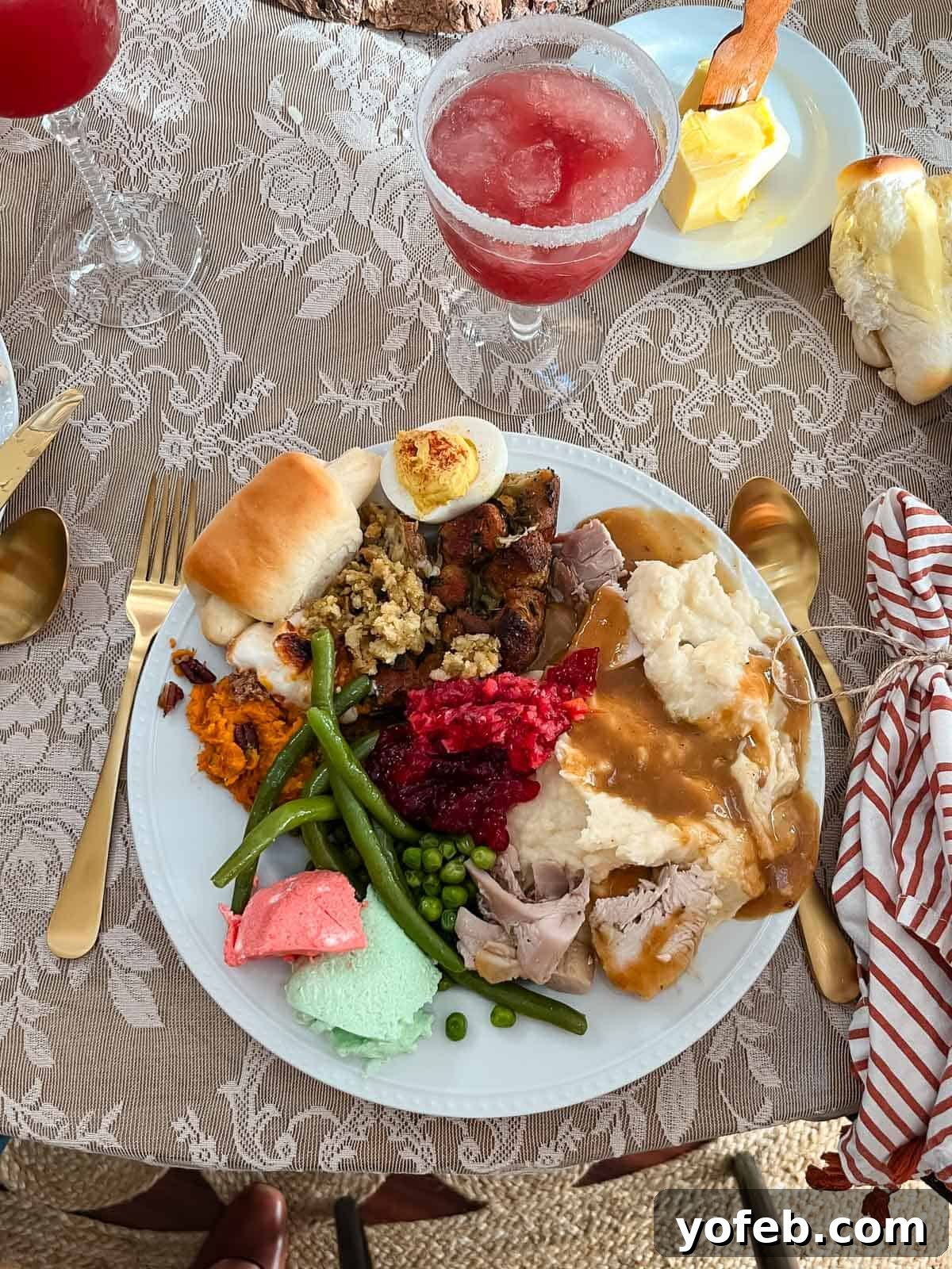 A plate filled with Thanksgiving food including turkey with gravy, mashed potatoes, stuffing, cranberry sauce, green beans, peas, sweet potatoes, a dinner roll, a deviled egg, and colorful desserts, with a pink drink beside it.