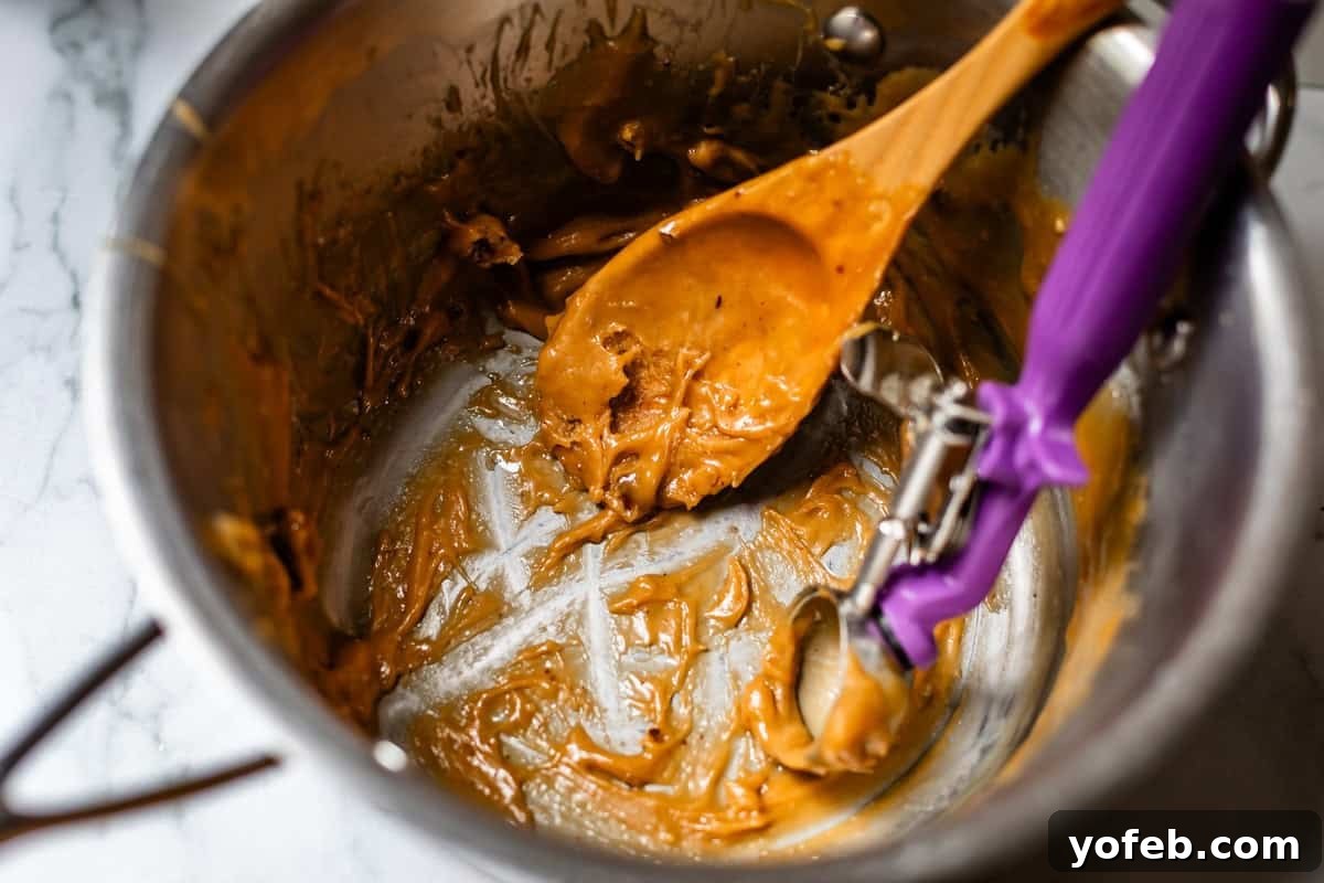 Buttery Pecan Bliss 20 A metal mixing bowl with remnants of thick, light brown batter, a wooden spoon, and a purple-handled cookie scoop, all resting on a white marble surface.
