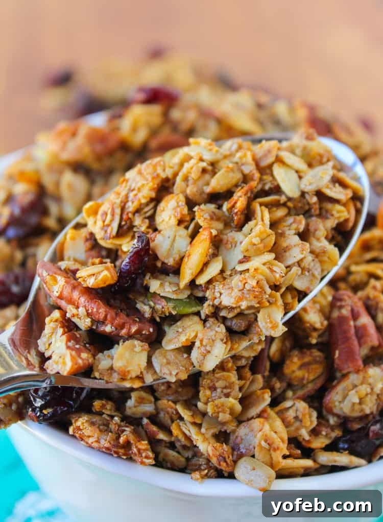 A spoonful of maple pecan granola with dried cherries held above a white bowl brimming with the wholesome breakfast treat.