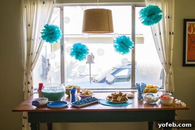 A table set for a Frozen birthday party with sandwiches, chips, snacks, a punch bowl, and plates. Blue paper pom-poms and white snowflake decorations hang in front of a sunny window with white curtains.