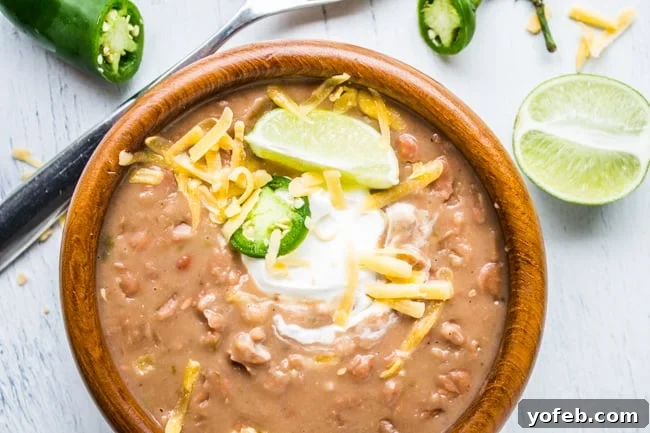 Traditional Tejano Pinto Beans (Slow Cooker) from The Food Charlatan. These Traditional Tejano Pinto Beans are the perfect blend of Southern and Mexican pinto beans styles. Tenderly slow cooked with a ham hock and jalapeños, the lime adds a little twist. Delicious as a side or perfect for a burrito! #pinto #beans #texan #crockpot #slowcooker #tejano #mexican #stovetop #recipes overhead shot of mexican pinto beans in a wooden bowl topped with cheese, jalapeño, sour cream, and lime.