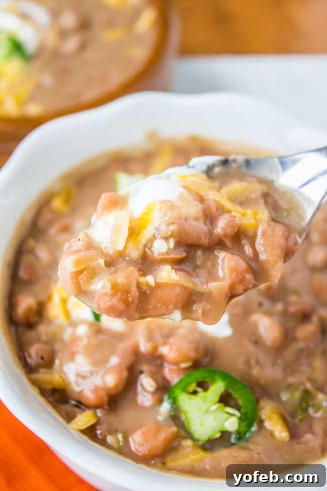 Classic Tejano Slow Cooker Frijoles 4 Mexican pinto beans recipe with sour cream, jalapeno, and cheddar being scooped up with a spoon.