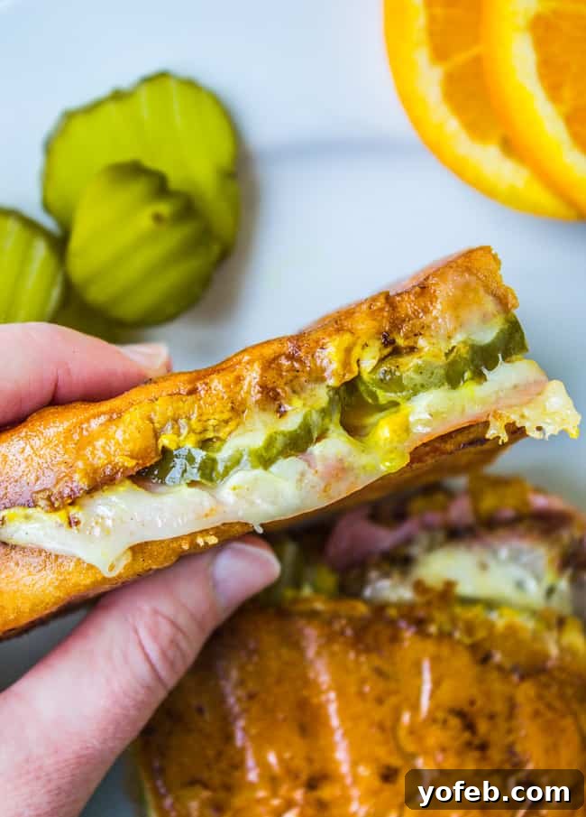 Pressed Perfection: The Ultimate Cubano 7 a hand holding a Cuban sandwich with pickles, cheese, pork, ham, and mustard.