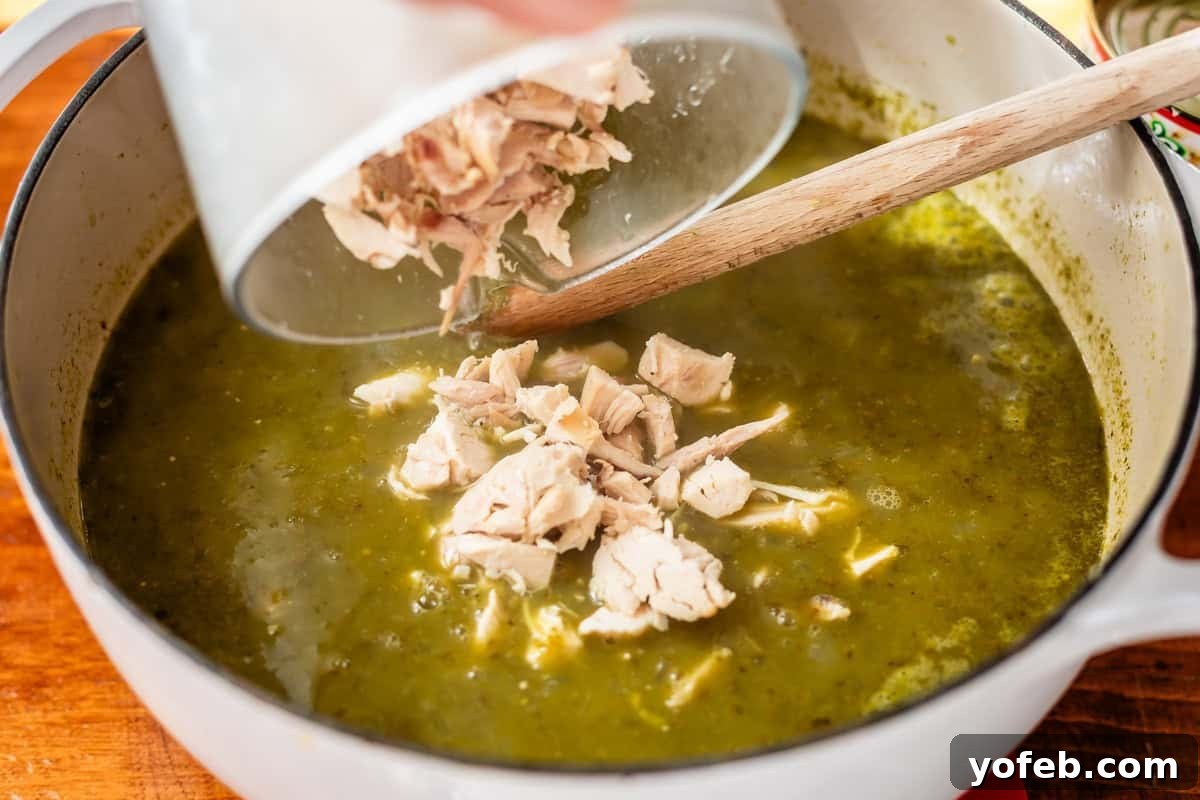 Vibrant Green Chicken Pozole in Minutes 20 A hand pours cooked, shredded chicken from a bowl into a pot of green soup with a wooden spoon inside, preparing a homemade meal.