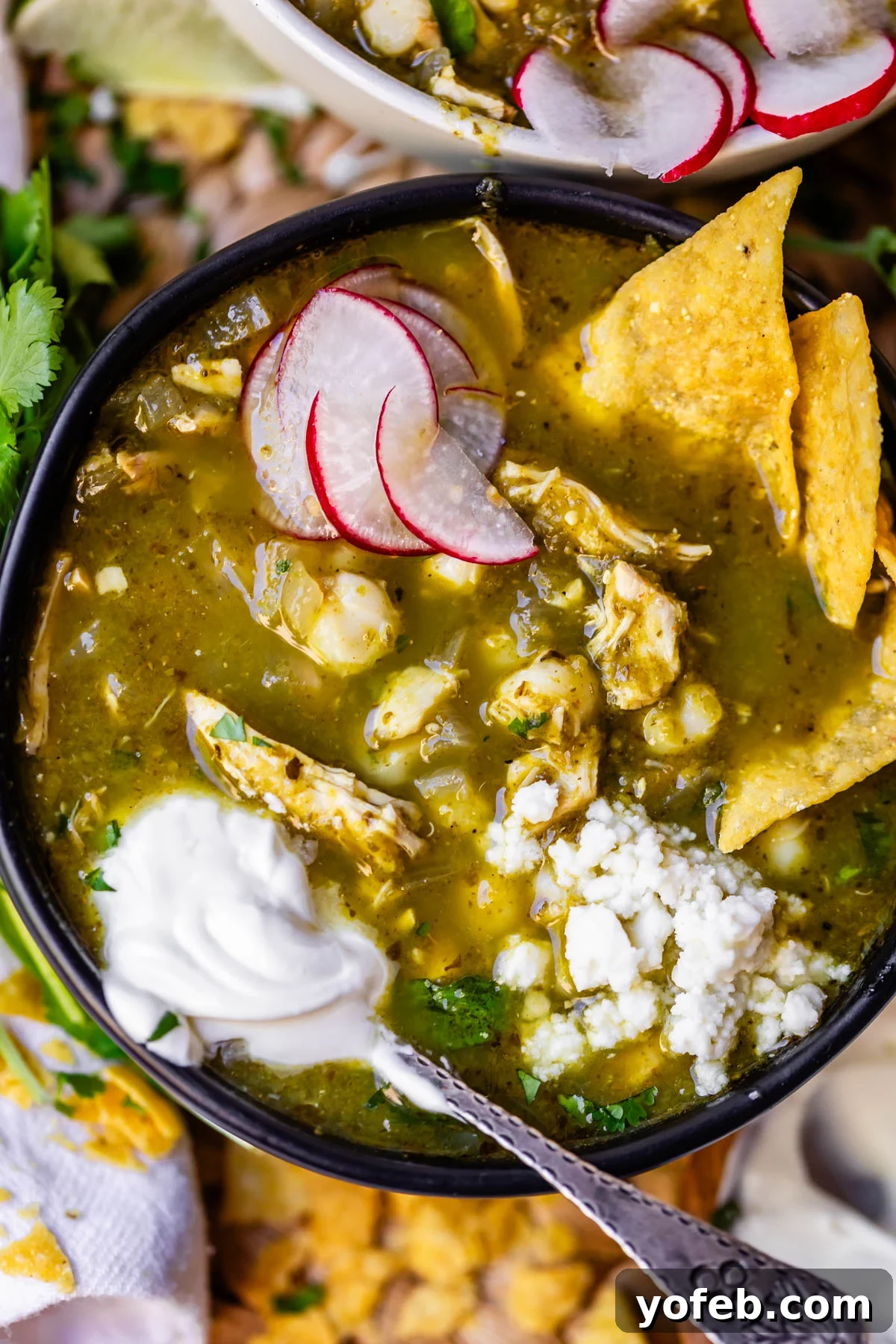 Vibrant Green Chicken Pozole in Minutes 22 A bowl of green chicken pozole topped with sliced radishes, crumbled cheese, tortilla chips, and a dollop of sour cream, with a spoon resting inside the bowl.