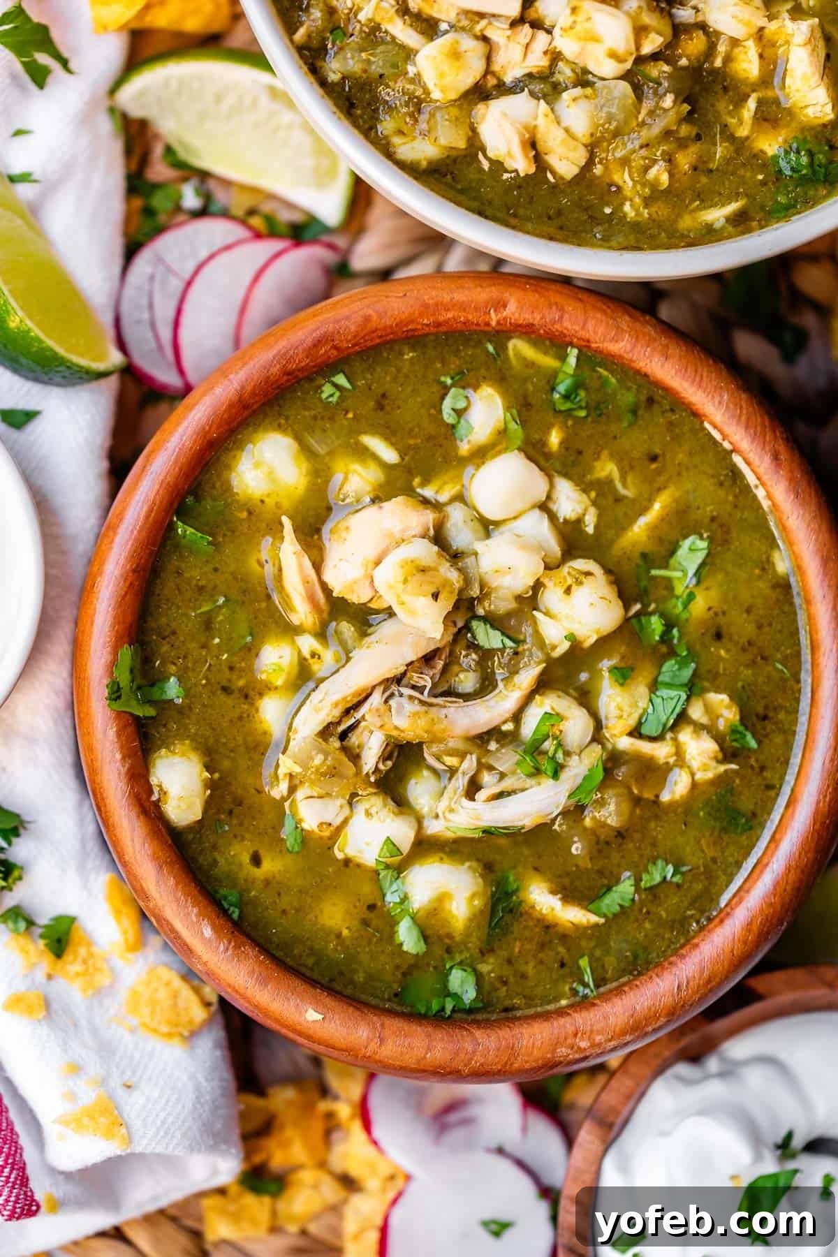 Vibrant Green Chicken Pozole in Minutes 26 A wooden bowl filled with green pozole soup, topped with shredded chicken, hominy, and chopped cilantro. Surrounding the bowl are lime wedges, sliced radishes, tortilla chips, and a small bowl of sour cream.
