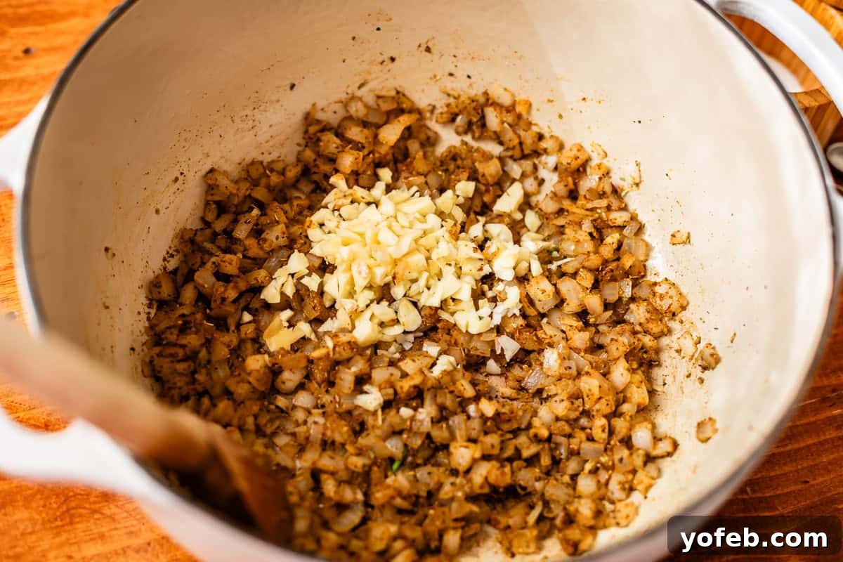 Vibrant Green Chicken Pozole in Minutes 9 Chopped onions and minced garlic are being sautéed with spices in a white pot, stirred with a wooden spoon, on a wooden surface.