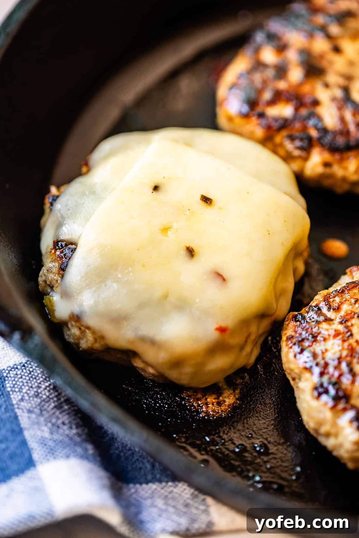 Game-Changing Turkey Burgers 11 A compelling close-up shot of a perfectly cooked burger patty, generously topped with glistening melted cheese, resting invitingly in a sleek black skillet. Adjacent to it, another plain patty awaits its cheesy crown. A corner of a soft blue and white cloth is subtly visible, adding a touch of rustic charm. This image highlights the crucial cheese melting step, promising a gooey, delicious outcome.