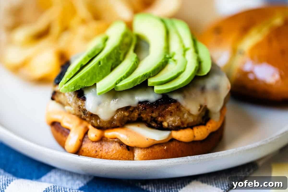 Game-Changing Turkey Burgers 12 A mouth-watering close-up of a perfectly grilled burger patty, generously topped with luscious melted cheese and creamy, fresh avocado slices. It rests invitingly on a beautifully toasted bun, complete with a swirl of savory sauce. The burger is presented elegantly on a plate, with a scattering of crispy potato chips blurred in the background, signaling a complete and satisfying meal.