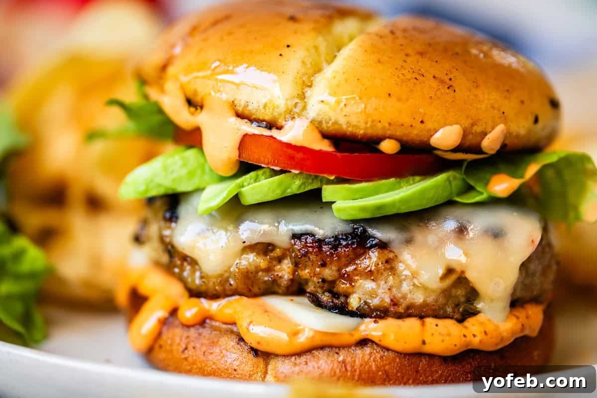 Game-Changing Turkey Burgers 13 A vibrant close-up of a perfectly crafted cheeseburger, featuring glistening melted cheese, crisp lettuce, ripe tomato, an abundance of creamy avocado slices, and a generous swirl of luscious sauce, all nestled within a beautifully toasted bun. In the background, a tantalizing blur of crispy fries completes the classic burger meal, showcasing the ultimate dining experience.