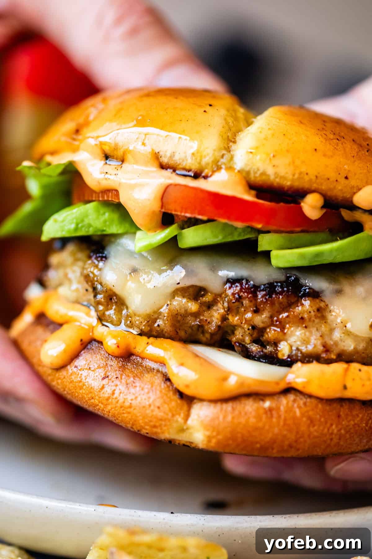 Game-Changing Turkey Burgers 15 A close-up of hands holding a magnificent cheeseburger, featuring perfectly melted cheese, crisp lettuce, ripe tomato, a luscious creamy sauce, and a glossy, toasted bun. The burger is incredibly juicy, with tantalizing sauce visibly dripping onto the plate below, a clear indication of its succulence and irresistible appeal.