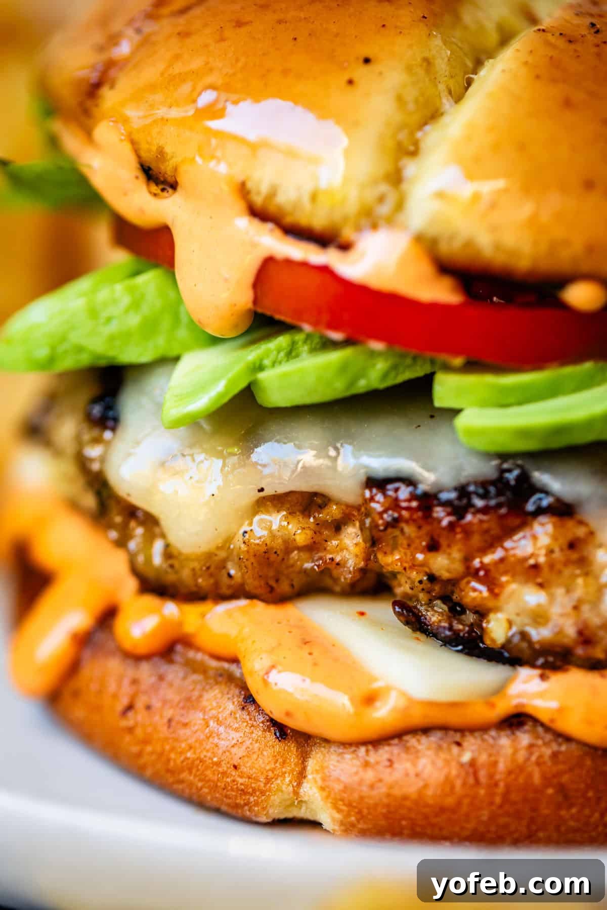 Game-Changing Turkey Burgers 17 A captivating close-up of a perfectly constructed turkey cheeseburger, featuring luscious melted cheese, vibrant slices of creamy avocado, ripe tomato, and a generous drizzle of creamy orange sauce, all artfully arranged on a golden, toasted bun. The burger is stacked impressively high, highlighting the freshness of its ingredients and the appealing texture of the sauces, signifying its deliciousness and the convenience of having it ready to enjoy any time.