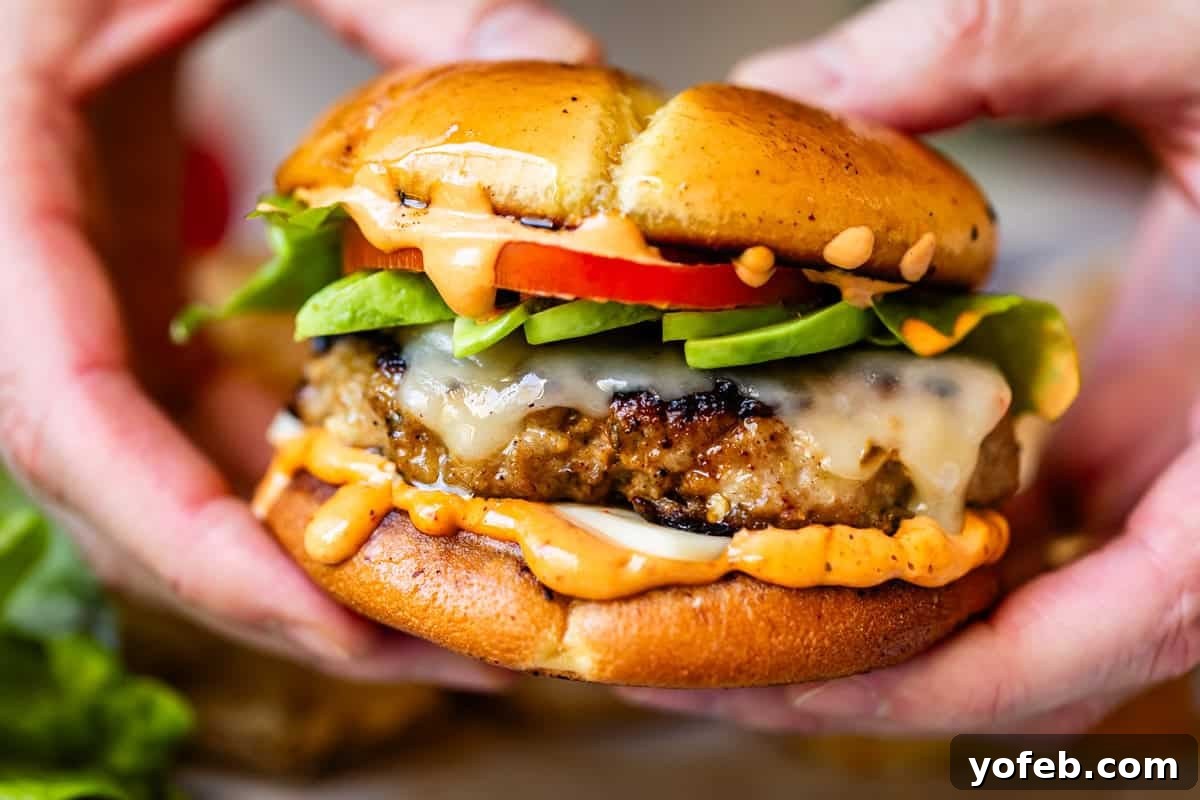 Game-Changing Turkey Burgers 5 Close-up of hands gently holding a juicy cheeseburger adorned with melted cheese, vibrant sliced avocado, crisp lettuce, ripe tomato, and a generous swirl of creamy sauce, all perfectly encased in a golden, toasted bun. The image effectively conveys the hands-on enjoyment and satisfying nature of the burger.