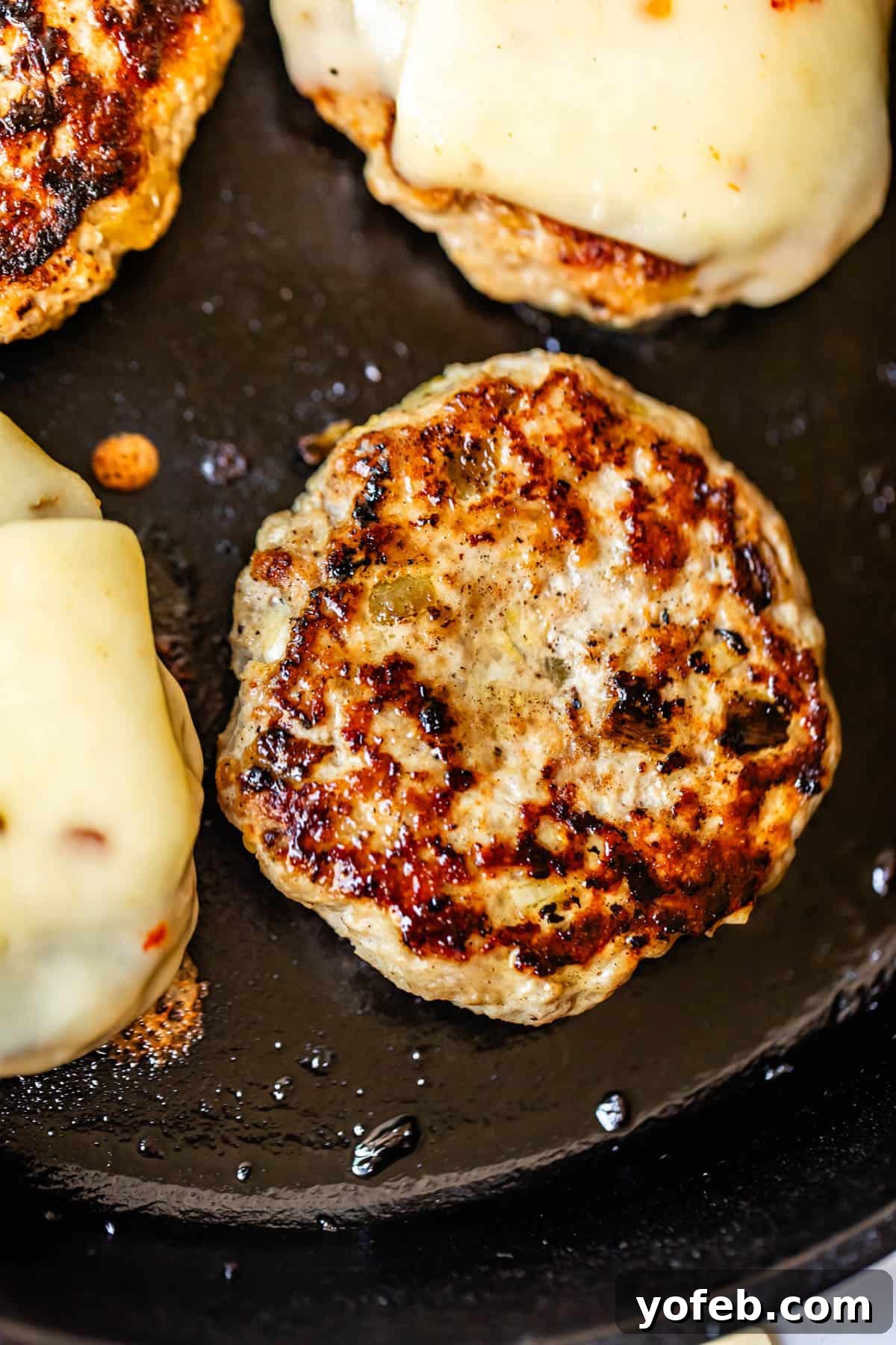 Game-Changing Turkey Burgers 10 A captivating close-up of several turkey burger patties sizzling and cooking beautifully in a hot skillet. Some of the patties are already topped with glorious melted cheese, and the surface of the pan reveals crispy, browned grill marks, indicating excellent searing and flavor development. This image vividly captures the cooking process and demonstrates the thumb divot in action, as the patties visibly flatten.