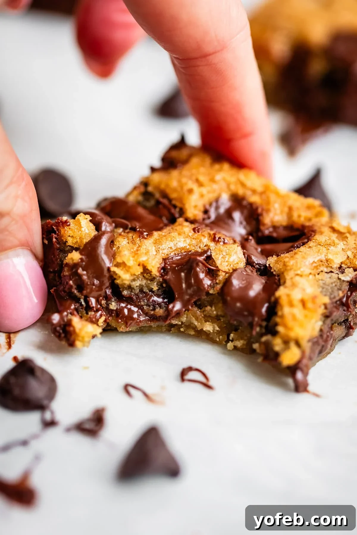 Chewy Chocolate Chip Squares 2 A hand holding a gooey, golden-brown chocolate chip cookie bar with melted chocolate chips, surrounded by chocolate chips on a white surface.