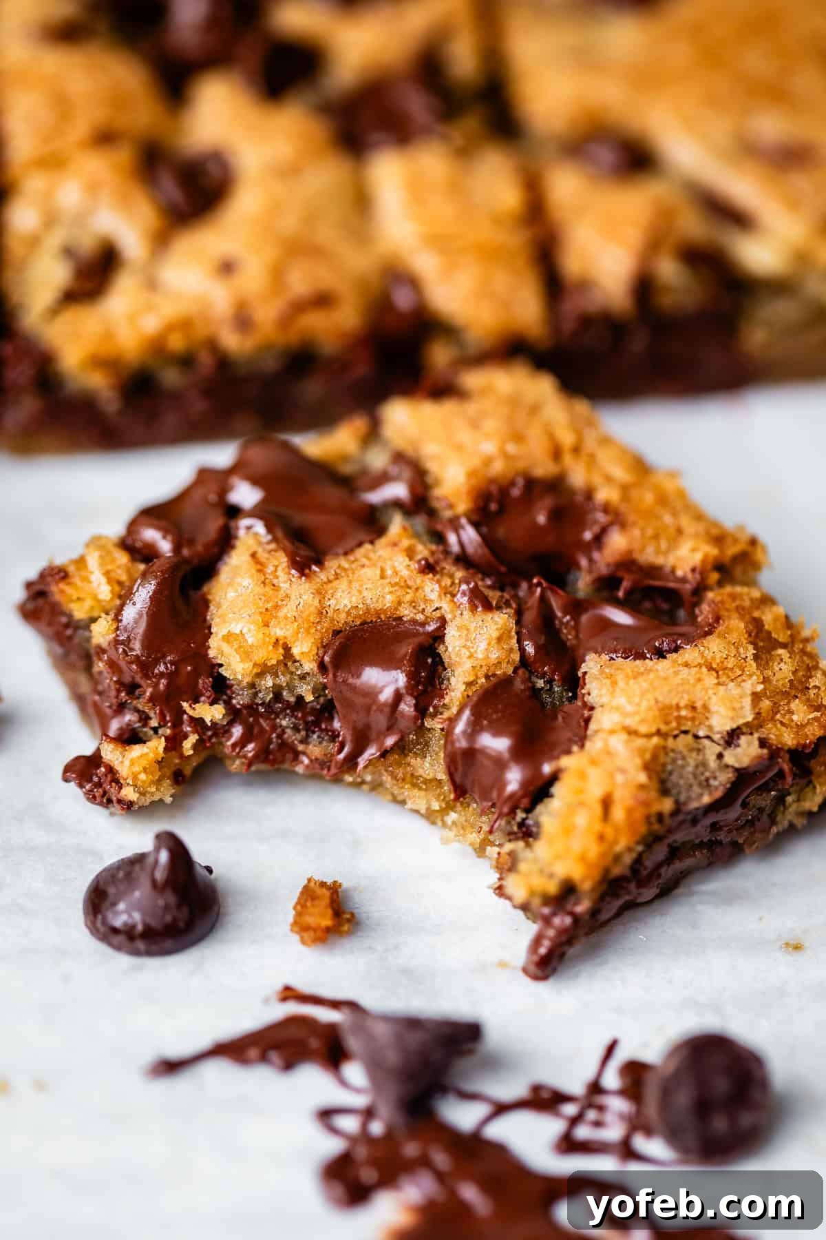 Chewy Chocolate Chip Squares 18 A close-up of a gooey chocolate chip cookie bar with melted chocolate chips, featuring a bite taken out. More bars and chocolate chips are visible in the background on a white surface.