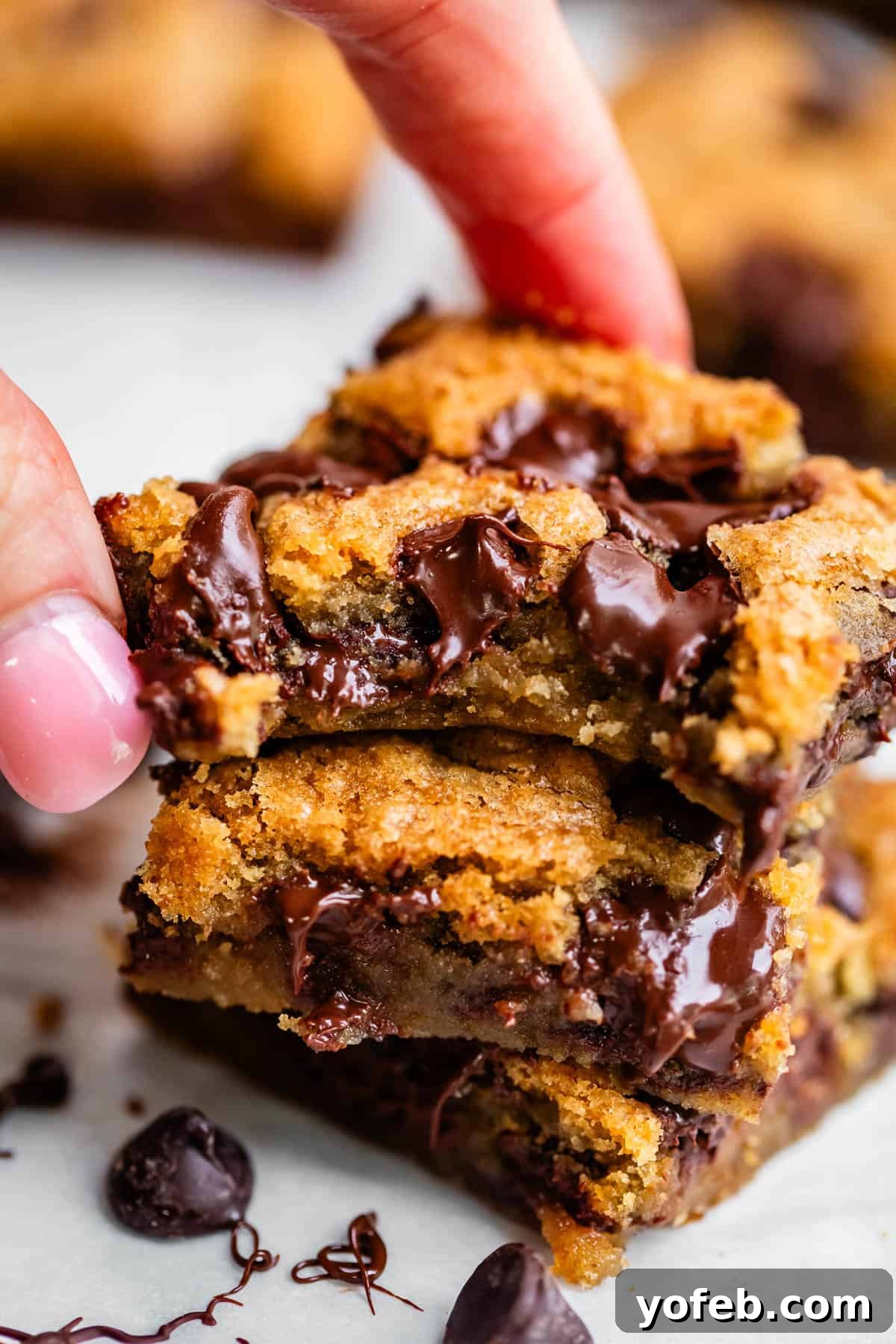 Chewy Chocolate Chip Squares 4 A close-up of a hand holding a stack of gooey chocolate chip cookie bars, with melted chocolate chips oozing from the soft, golden-brown centers.