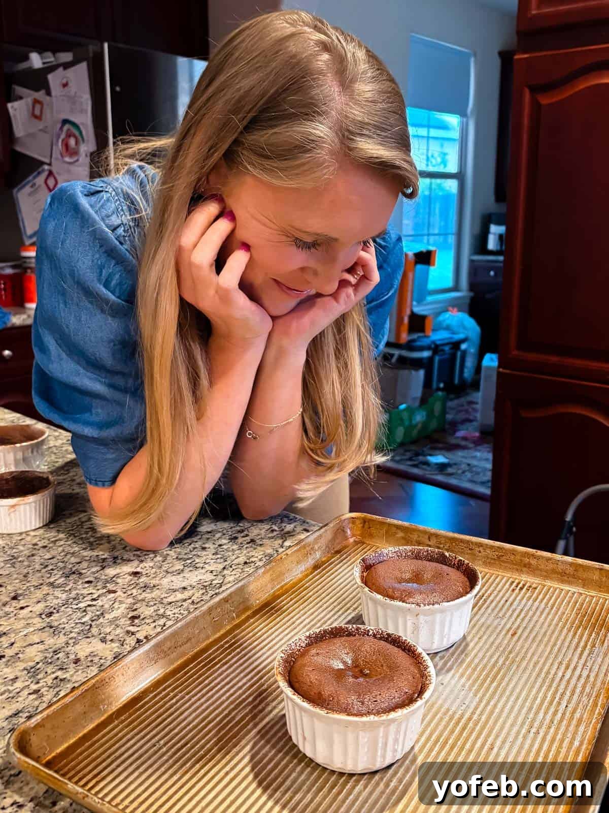 Molten Chocolate Magic 24 A woman with blonde hair leans over a kitchen counter, smiling and looking at two chocolate soufflés in white ramekins on a baking tray. The kitchen has wooden cabinets and a refrigerator in the background.