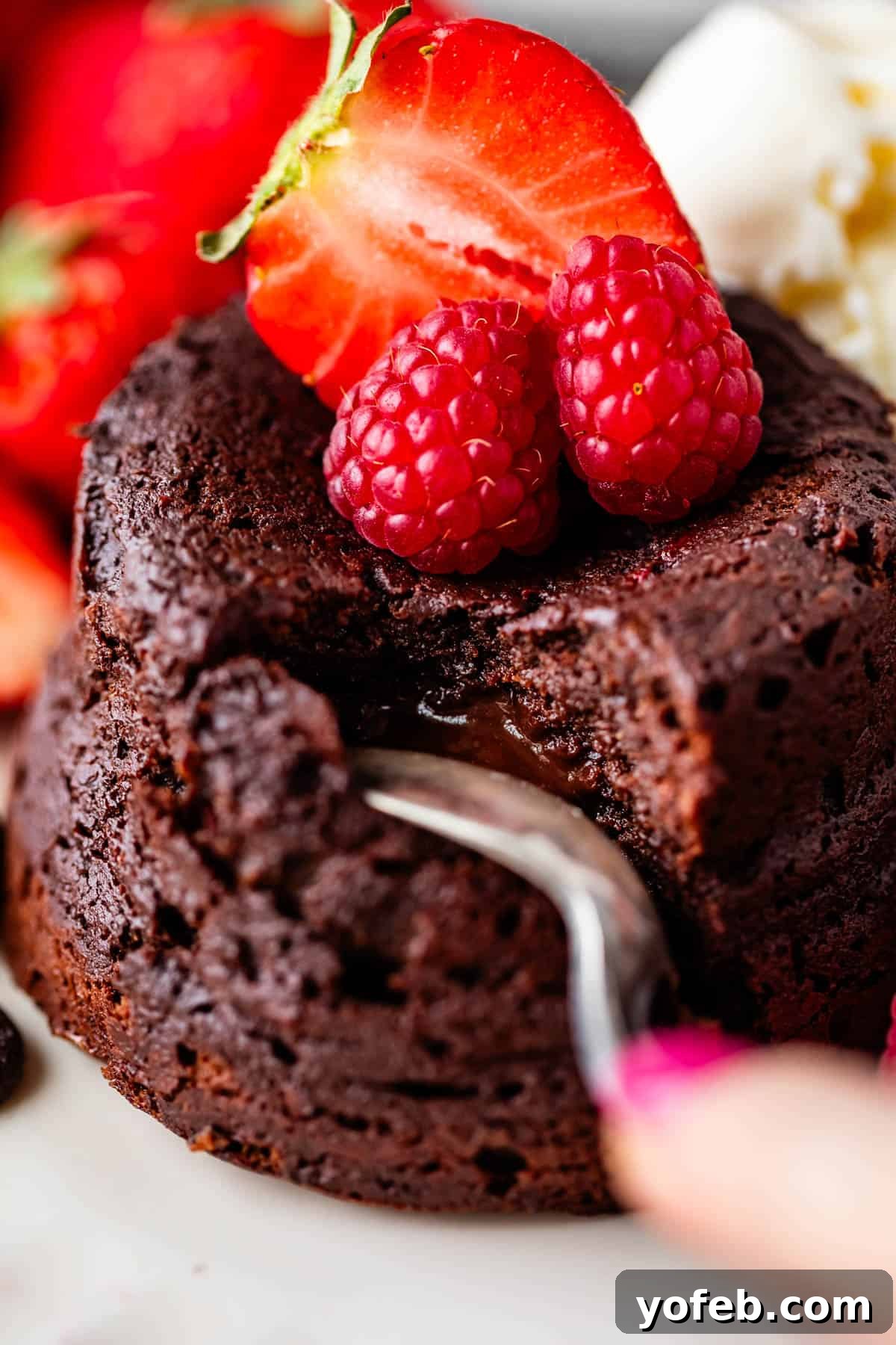 Molten Chocolate Magic 4 Close-up of a chocolate lava cake with a molten center, topped with fresh raspberries and a halved strawberry. A fork is breaking into the cake. In the background, there are more strawberries and a scoop of vanilla ice cream.