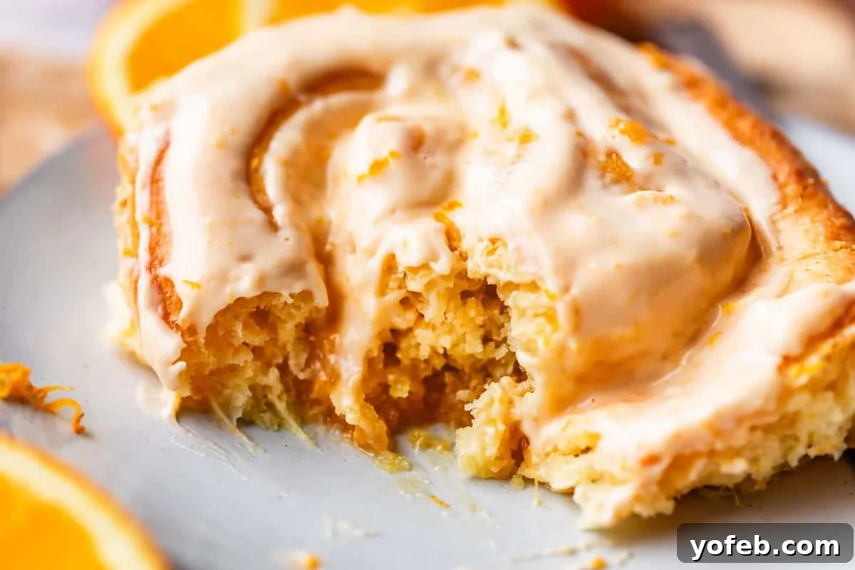 Zesty Orange Sweet Rolls 29 A delectable close-up of a partially eaten orange cinnamon roll, artfully presented on a light-colored plate. The roll is generously topped with creamy icing and delicate orange zest, while fresh orange slices blur softly in the background, completing an appetizing scene.