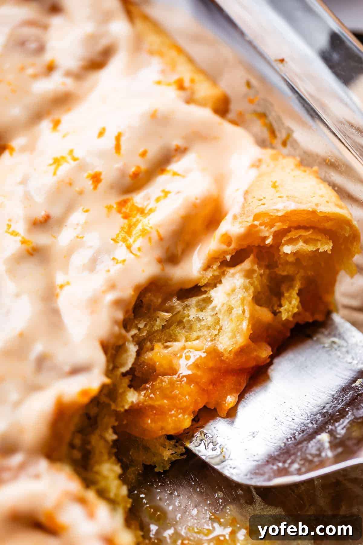 Zesty Orange Sweet Rolls 30 A close-up shot captures a glazed pastry being lifted with a gleaming metal spatula, revealing its irresistible gooey orange filling. The pastry is crowned with a luscious, cream-colored icing and sprinkled with vibrant orange zest, showcasing its delightful texture and rich flavor.
