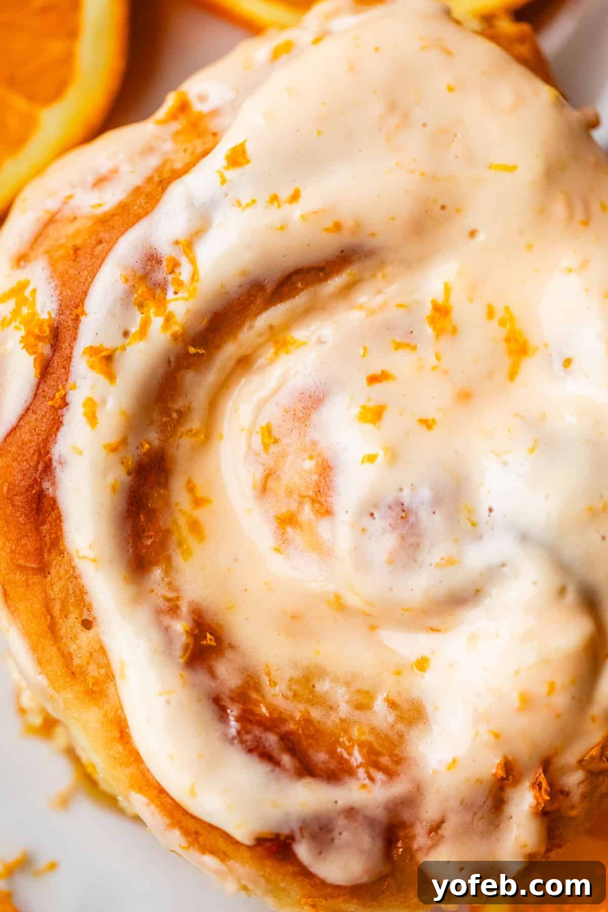 Zesty Orange Sweet Rolls 4 A decadent close-up of a freshly baked orange roll, generously coated with a rich, creamy orange icing and delicately sprinkled with bright orange zest. A blurred slice of fresh orange sits elegantly in the background, enhancing the citrus theme.