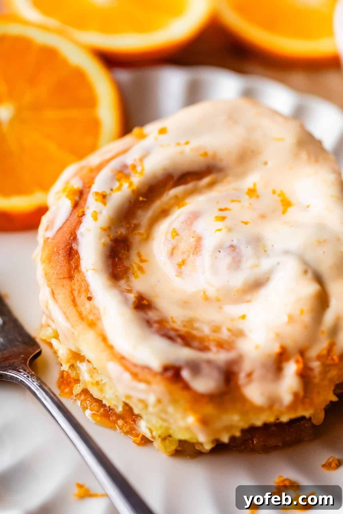 Zesty Orange Sweet Rolls 31 A delightful close-up of an orange sweet roll, beautifully topped with creamy icing and vibrant orange zest, presented on a pristine white plate. A silver fork rests beside it, inviting a taste, with fresh orange slices blurred elegantly in the background, completing a tempting breakfast scene.