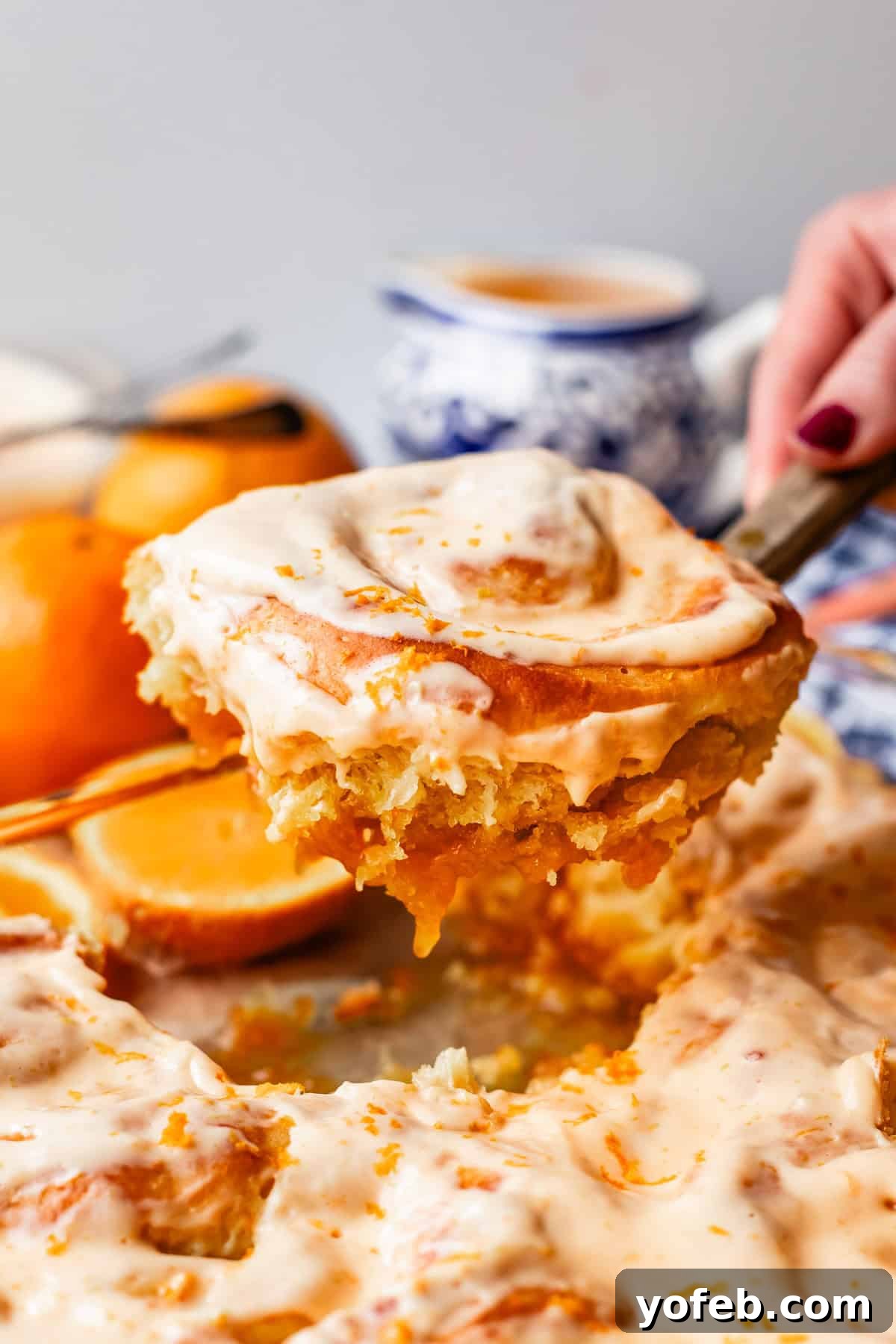 Zesty Orange Sweet Rolls 33 A hand gracefully lifts a freshly frosted cinnamon roll, generously topped with vibrant orange zest, from a tray filled with similar irresistible rolls. In the softly blurred background, an arrangement of fresh oranges, a ceramic cup, and a charming blue-and-white teapot complete the inviting breakfast tableau.