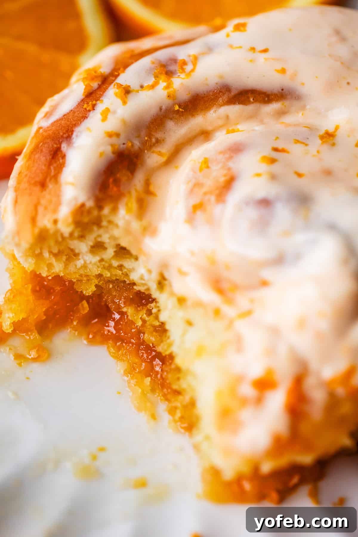 Zesty Orange Sweet Rolls 5 A tantalizing close-up of a partially eaten orange roll, showcasing its fluffy texture and rich, gooey orange filling. The roll is topped with a generous swirl of creamy orange glaze and bright orange zest. In the softly blurred background, vibrant orange slices complete the inviting culinary scene.