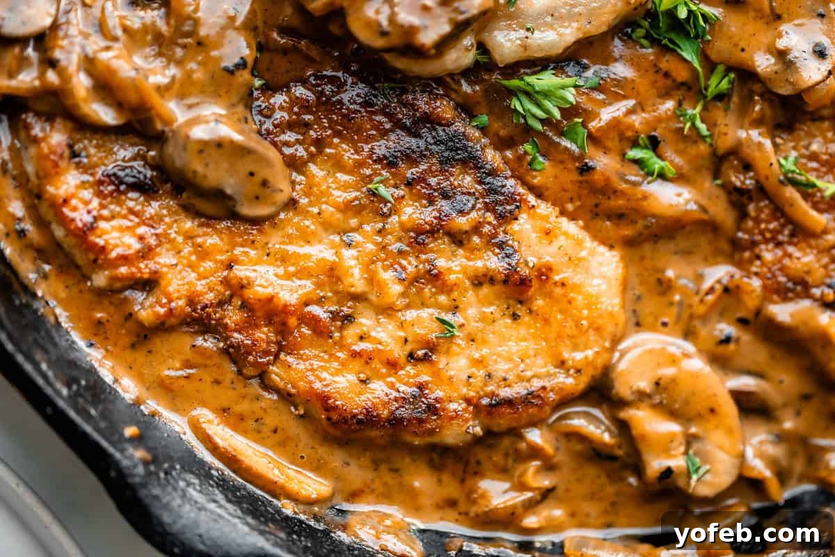 Tender Smothered Pork Chops 20 Close-up of a skillet with golden-brown, seared pork chops in a creamy mushroom sauce, garnished with chopped parsley.