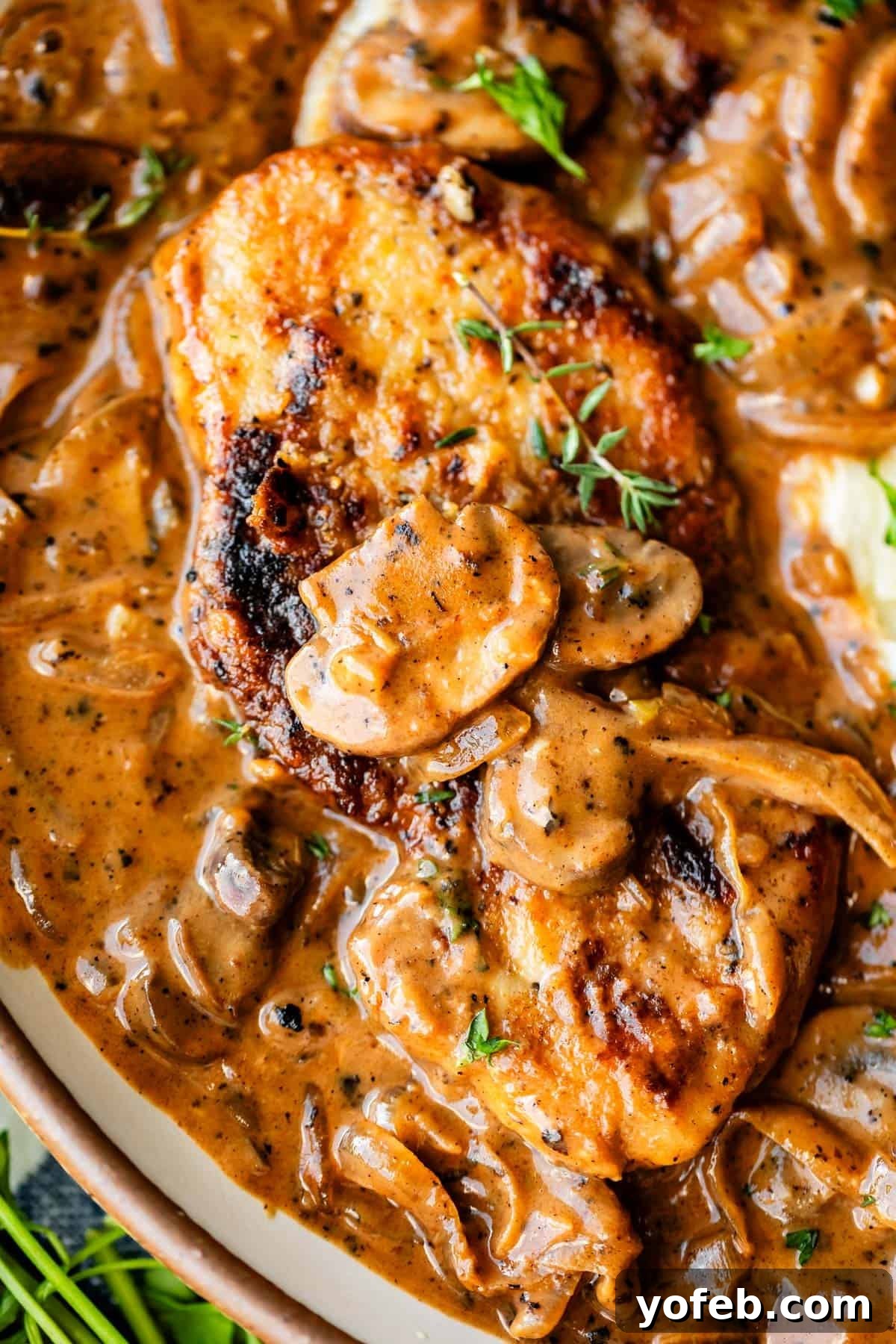 Tender Smothered Pork Chops 3 A close-up of a juicy, golden-brown grilled chicken breast topped with sliced mushrooms and herbs, covered in a creamy brown sauce with onions and garnished with a sprig of fresh thyme.