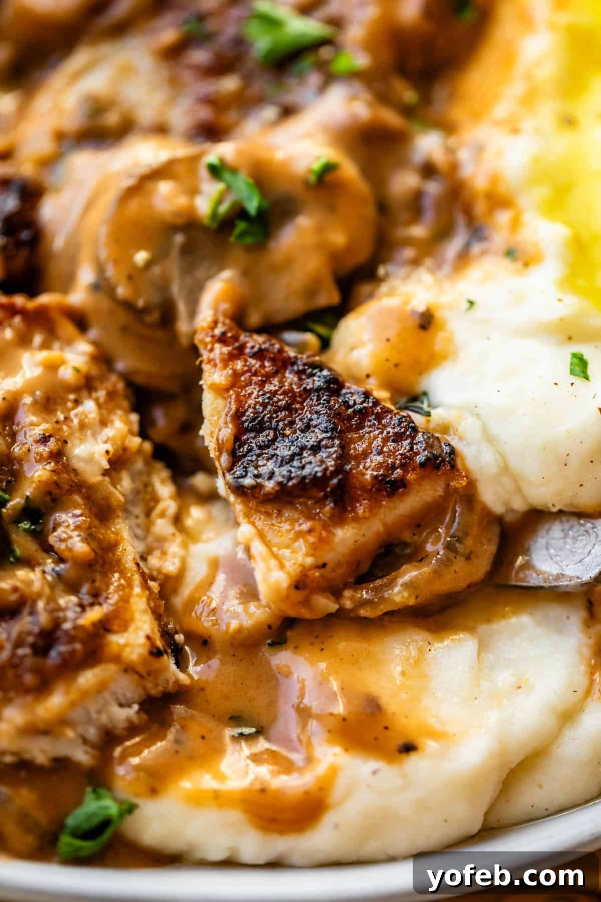 Tender Smothered Pork Chops 21 Close-up of grilled chicken pieces covered in creamy mushroom sauce, served on a bed of mashed potatoes with herbs sprinkled on top.